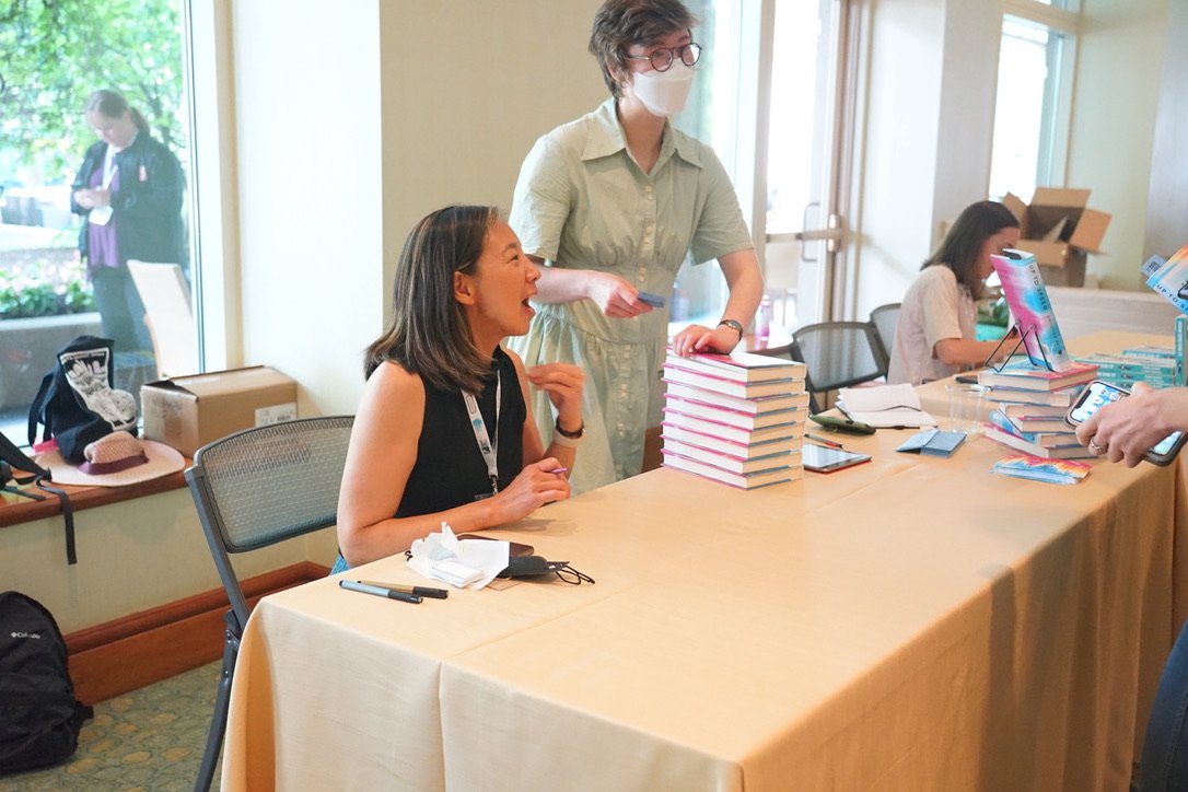 A woman seated at a table at a book signing event, smiling and engaging with a person holding a book. A woman standing behind the table, wearing a face mask, is arranging books. There are stacks of books on the table and another person working at a c