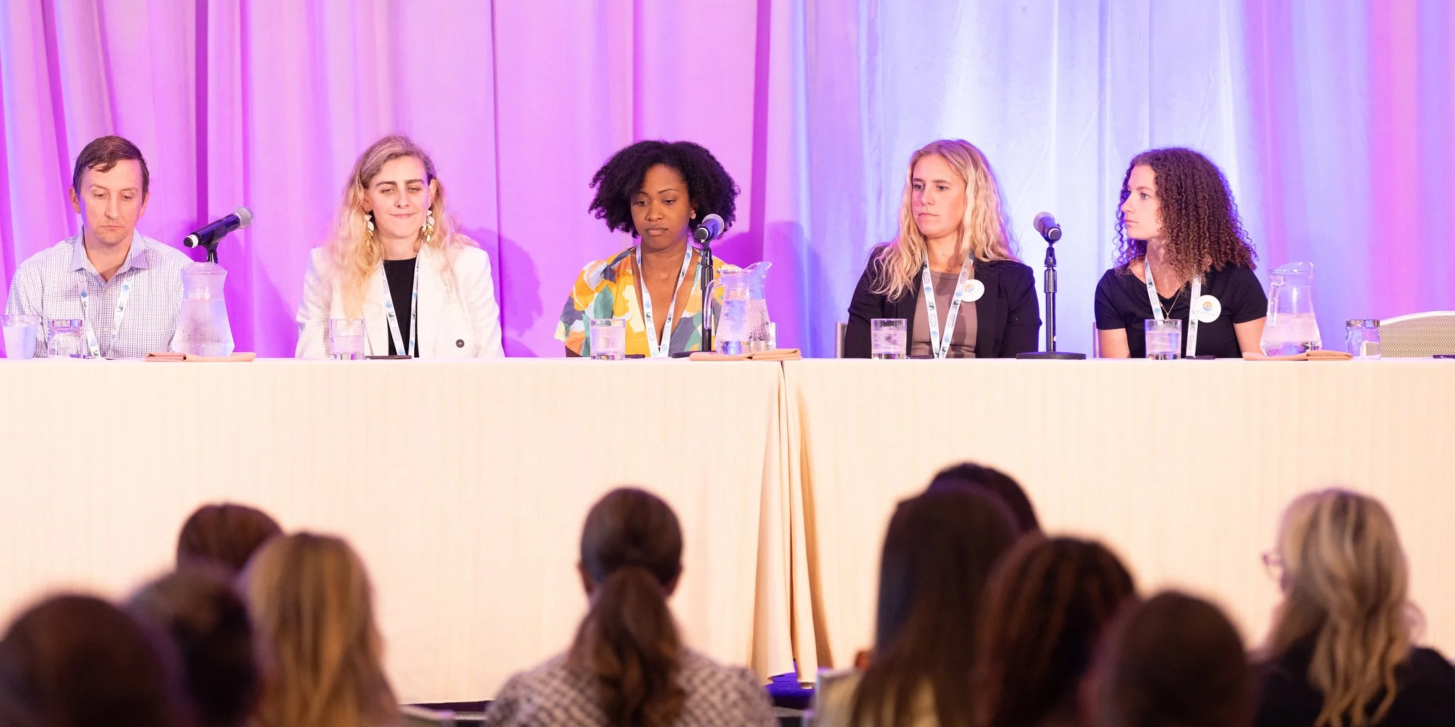 Five women sit at a long table on a stage, participating in a panel discussion. They are facing an audience, with purple and white curtains in the background. There are microphones and water pitchers on the table.