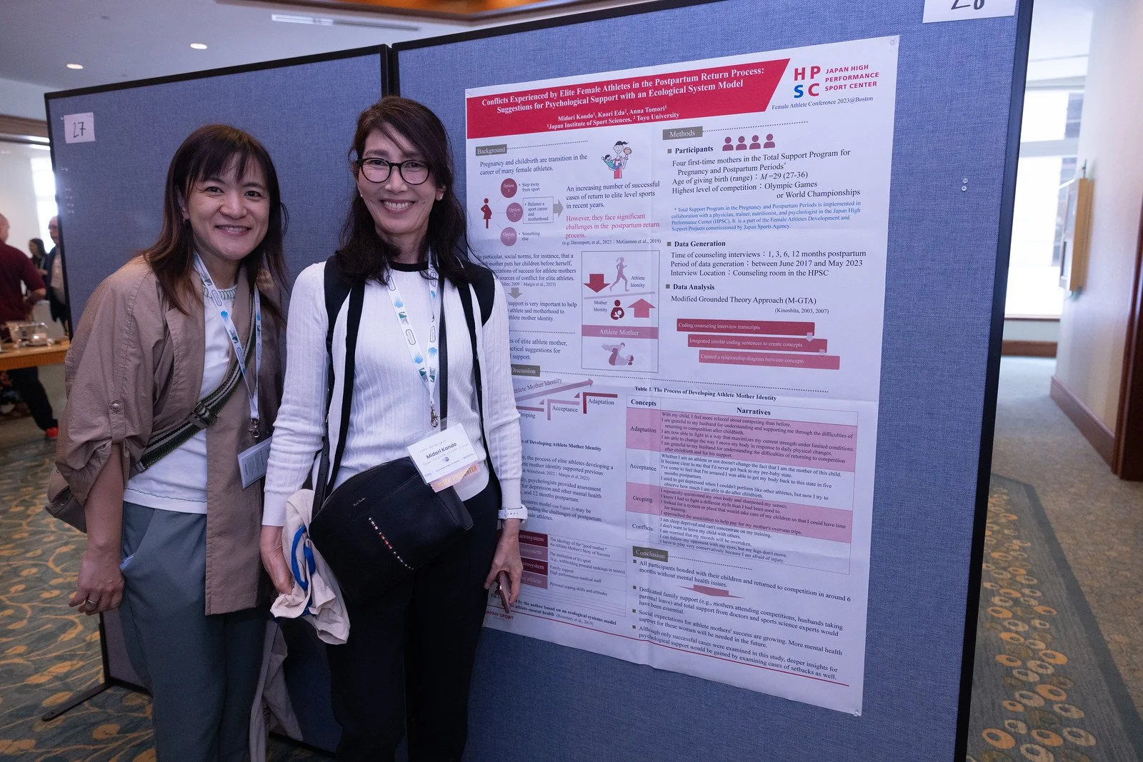 Two women standing next to a scientific research poster at a conference exhibition, smiling at the camera.