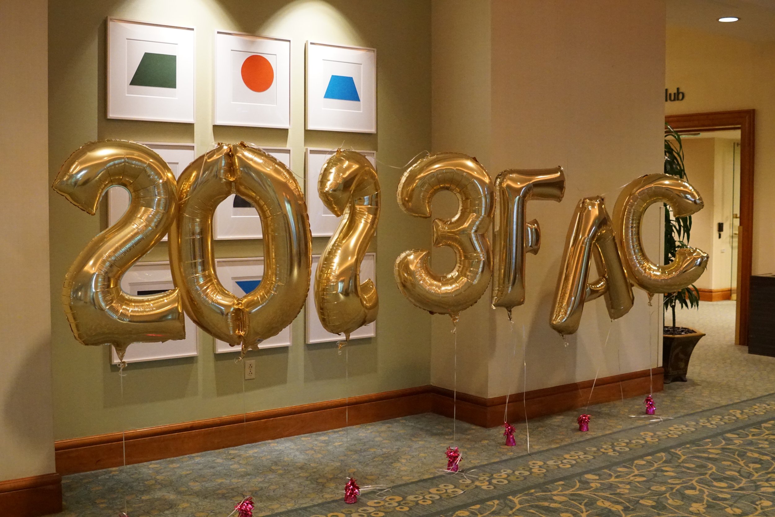 Gold foil balloons spelling out '2023' on a decorated wall with framed abstract art and a doorway in the background.