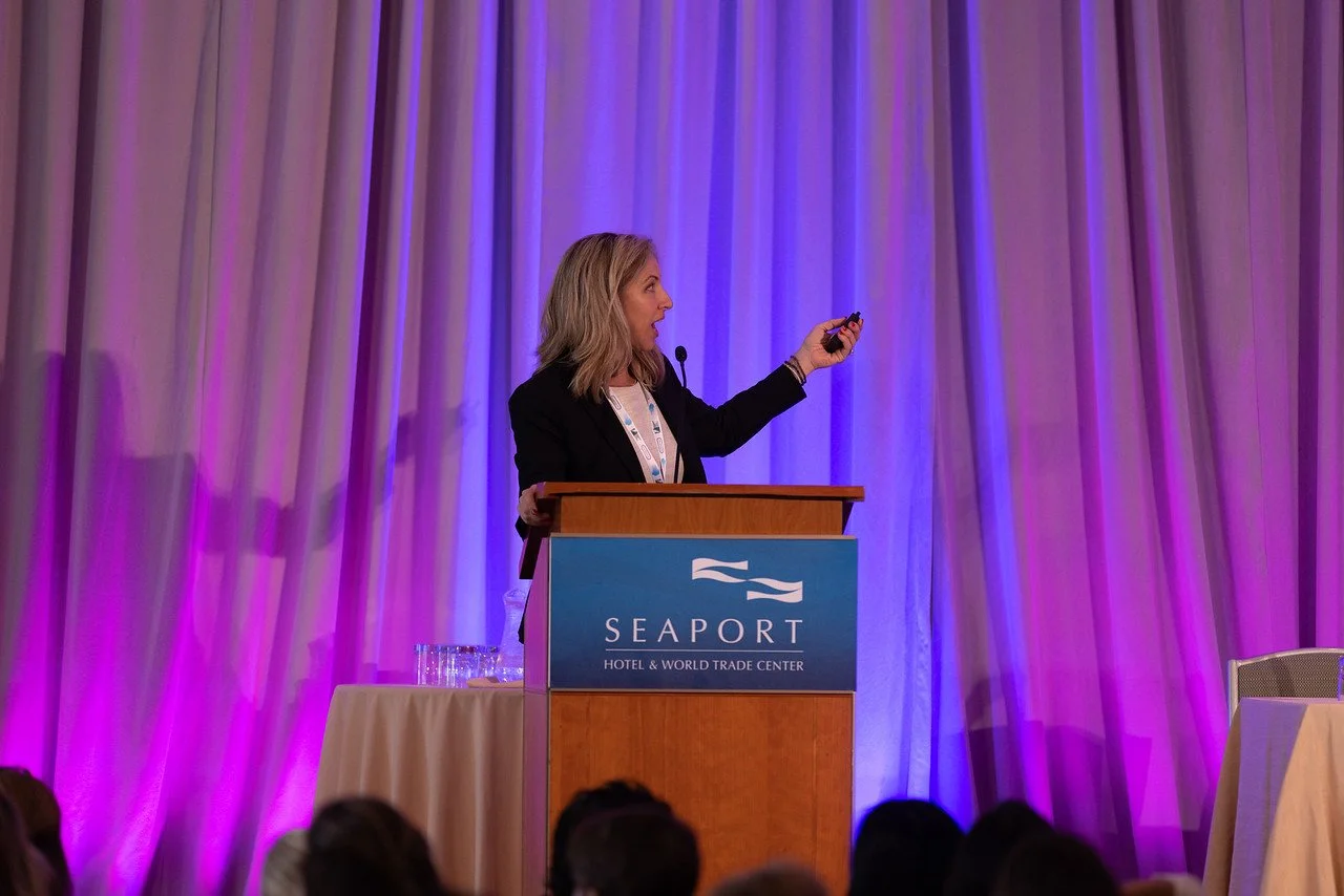 A woman giving a presentation at a podium with a sign that says "Seaport Hotel & World Trade Center," in front of purple and blue curtains, with a few glasses of water on a table beside her.