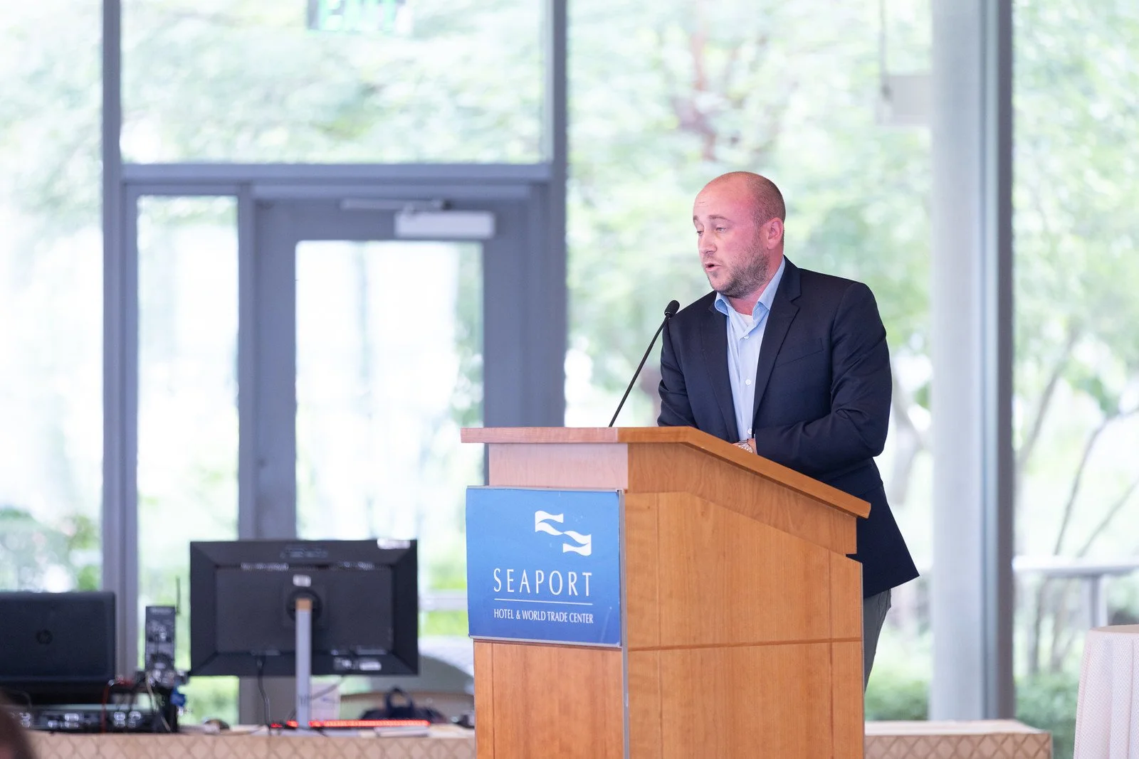 A man in a dark suit speaks at a podium with a blue sign that reads 'SEAPORT HOTEL & WORLD TRADE CENTER'. There are computer monitors and electronic equipment in front of him, and large windows with a view of greenery in the background.