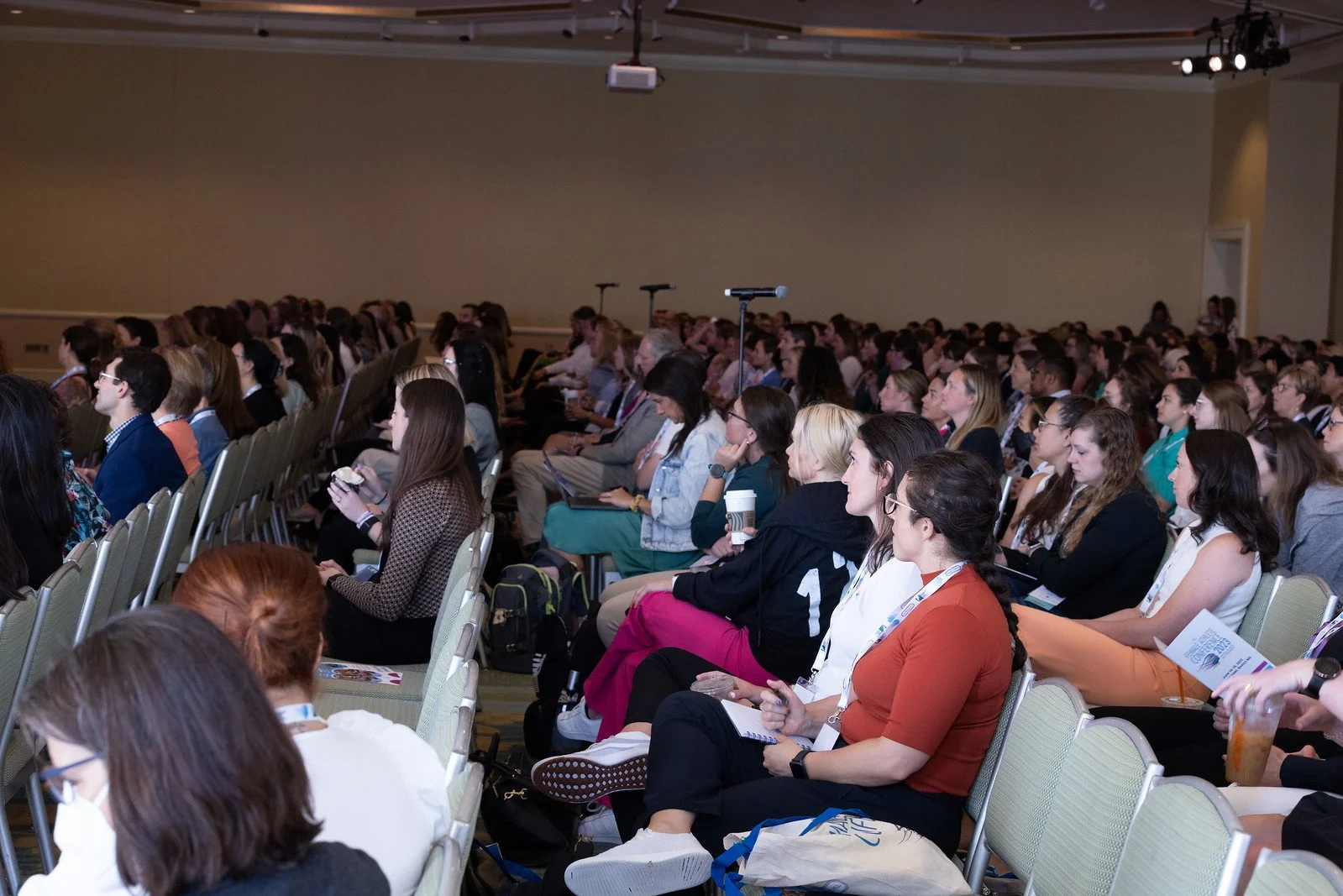 Audience attending a conference or seminar in a large hall, seated in rows, listening to a speaker or presentation.