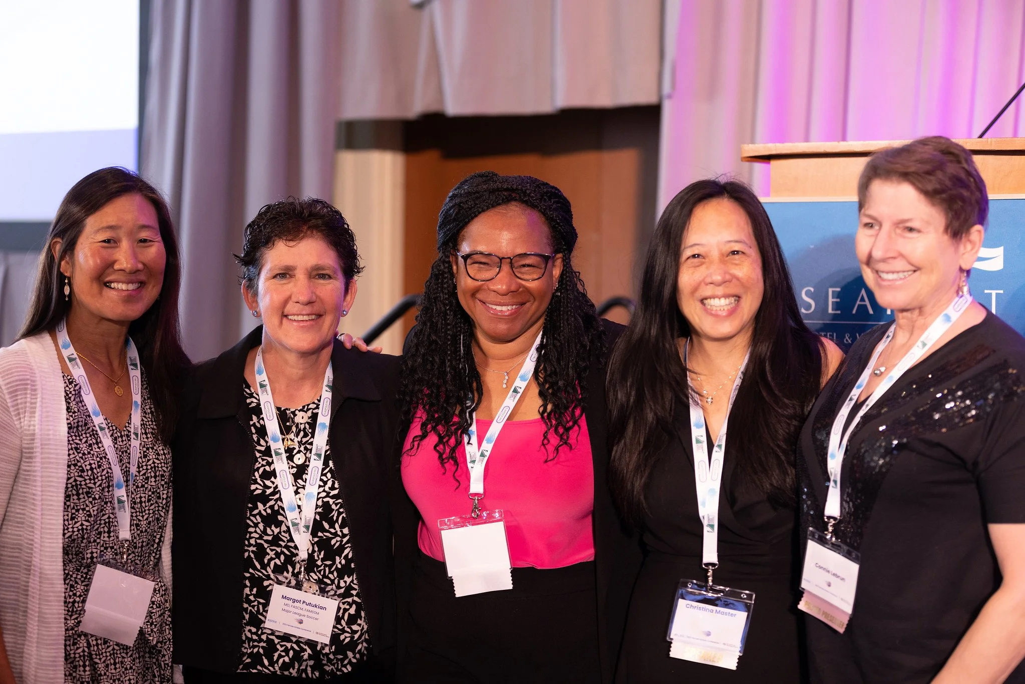 A group of five diverse women smiling at a conference with name tags around their necks.