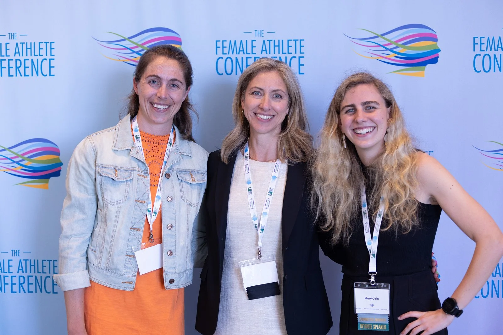 Three women smiling at the Female Athlete Conference, standing in front of a backdrop with the event logo. They are wearing conference badges around their necks.