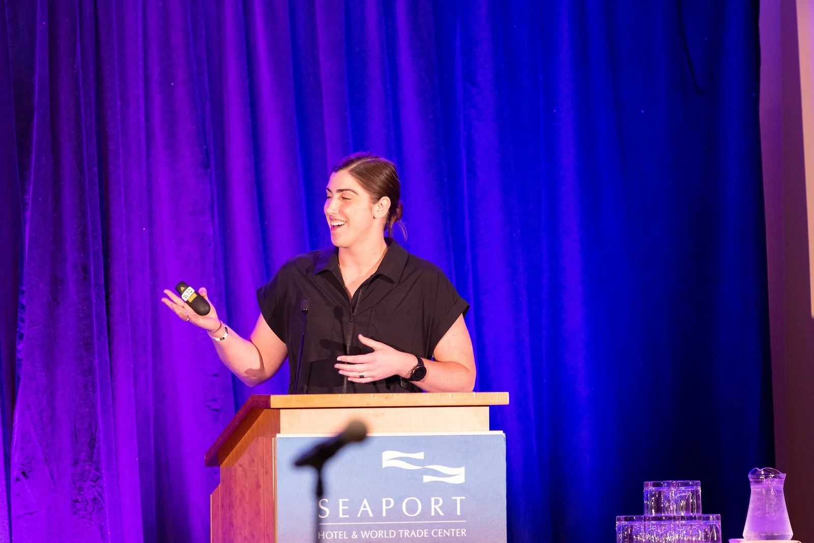 A woman speaking at a podium with a blue backdrop and purple curtains at Seaport Hotel & World Trade Center, holding a wireless presentation device, smiling.