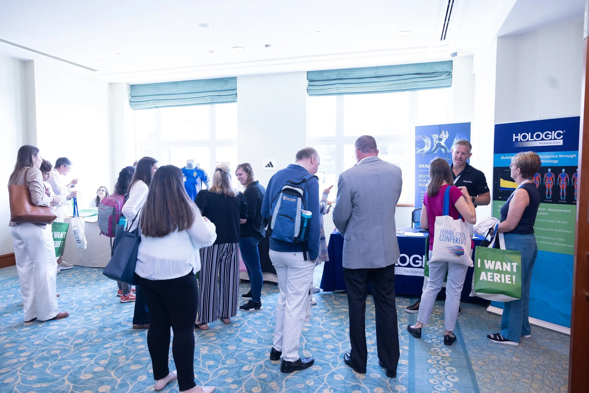 People attending a conference or expo in a brightly lit room with large windows. Some are gathered around exhibitor booths, engaging in conversations and looking at displays. There are banners and posters related to scientific or medical technology, 