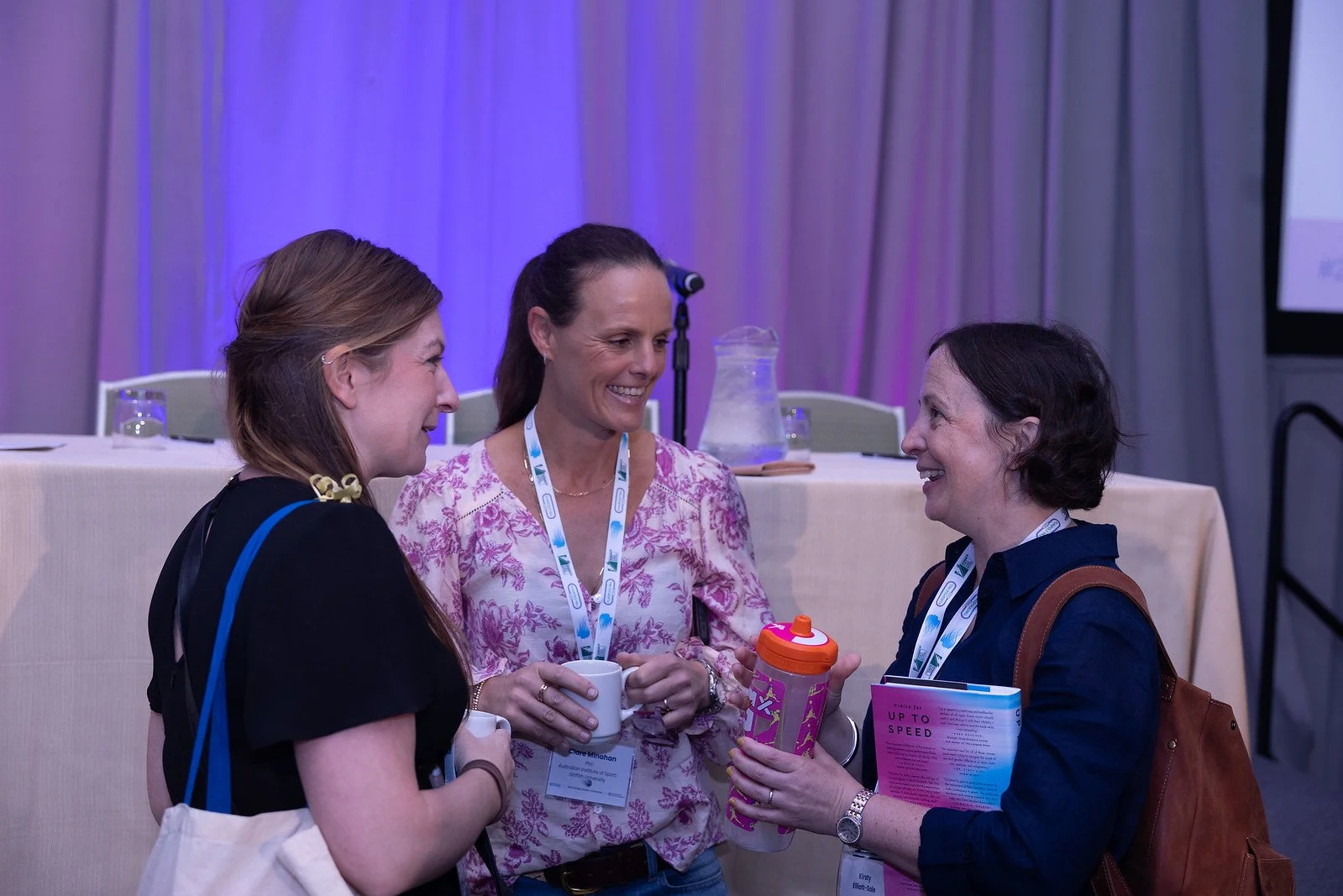 Three women at a conference having a conversation, holding drinks and a children's sippy cup, with conference badges hanging around their necks.