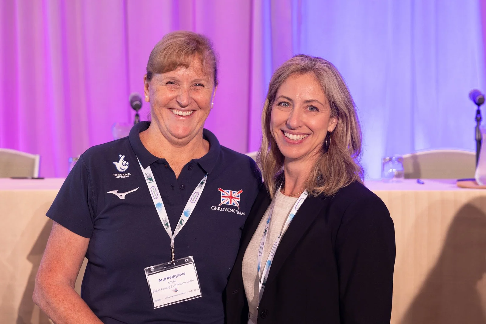 Two women smiling at an indoor conference or event. The woman on the left wears a navy polo with logos, and the woman on the right wears a black blazer. Both have conference badges on lanyards around their necks.