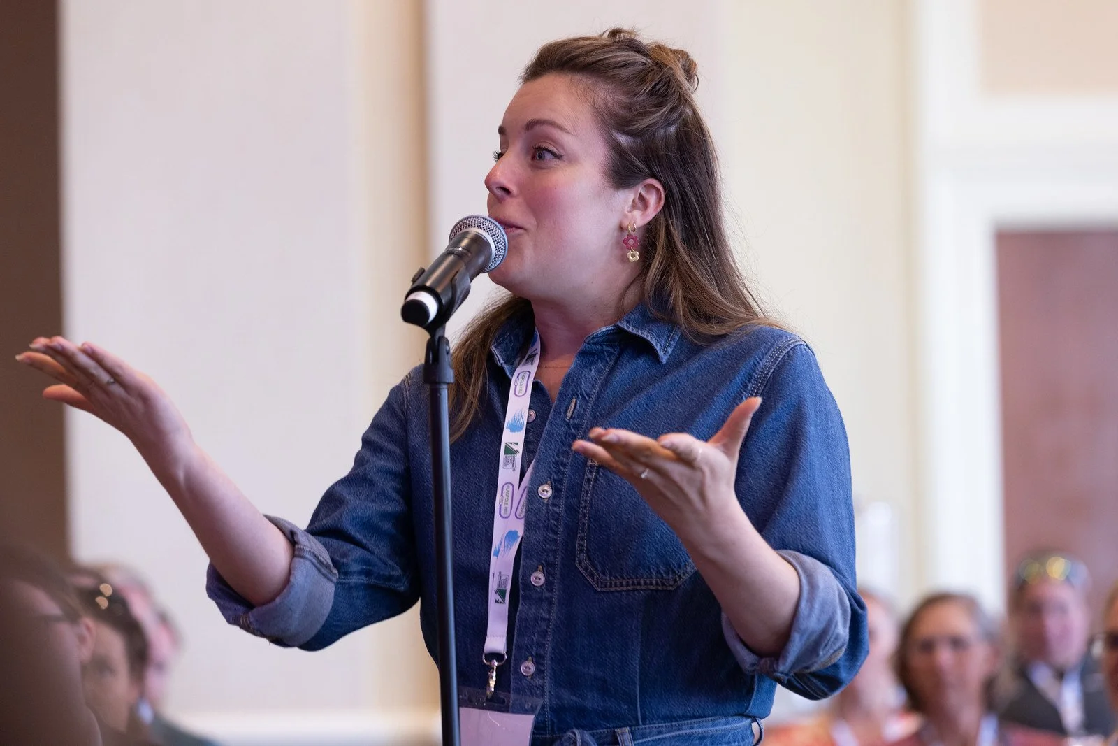 A woman with brown hair dressed in a denim shirt speaks into a microphone during a conference or seminar, gesturing with her hands, with a group of women seated in the background.