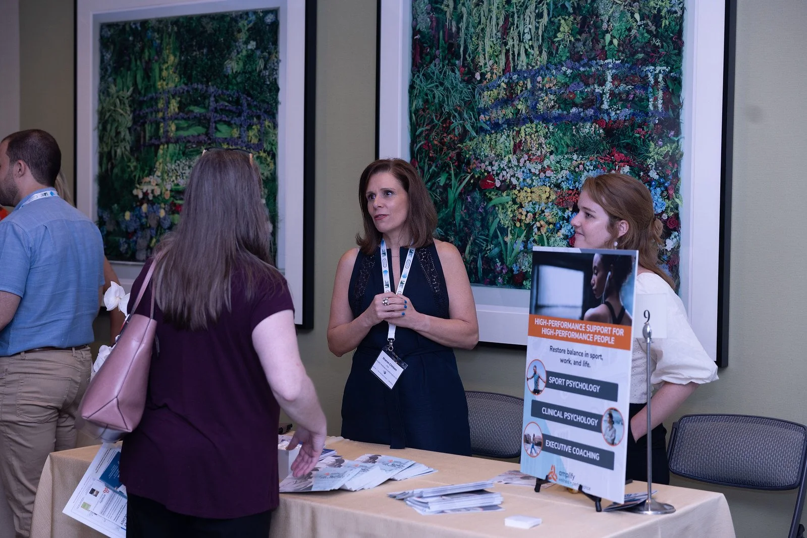 Three women are at a conference, engaged in conversation. One woman is wearing a navy dress with a lanyard, another has a maroon shirt and a pink purse, and the third is in a white shirt. They are standing next to a table with brochures and a sign pr