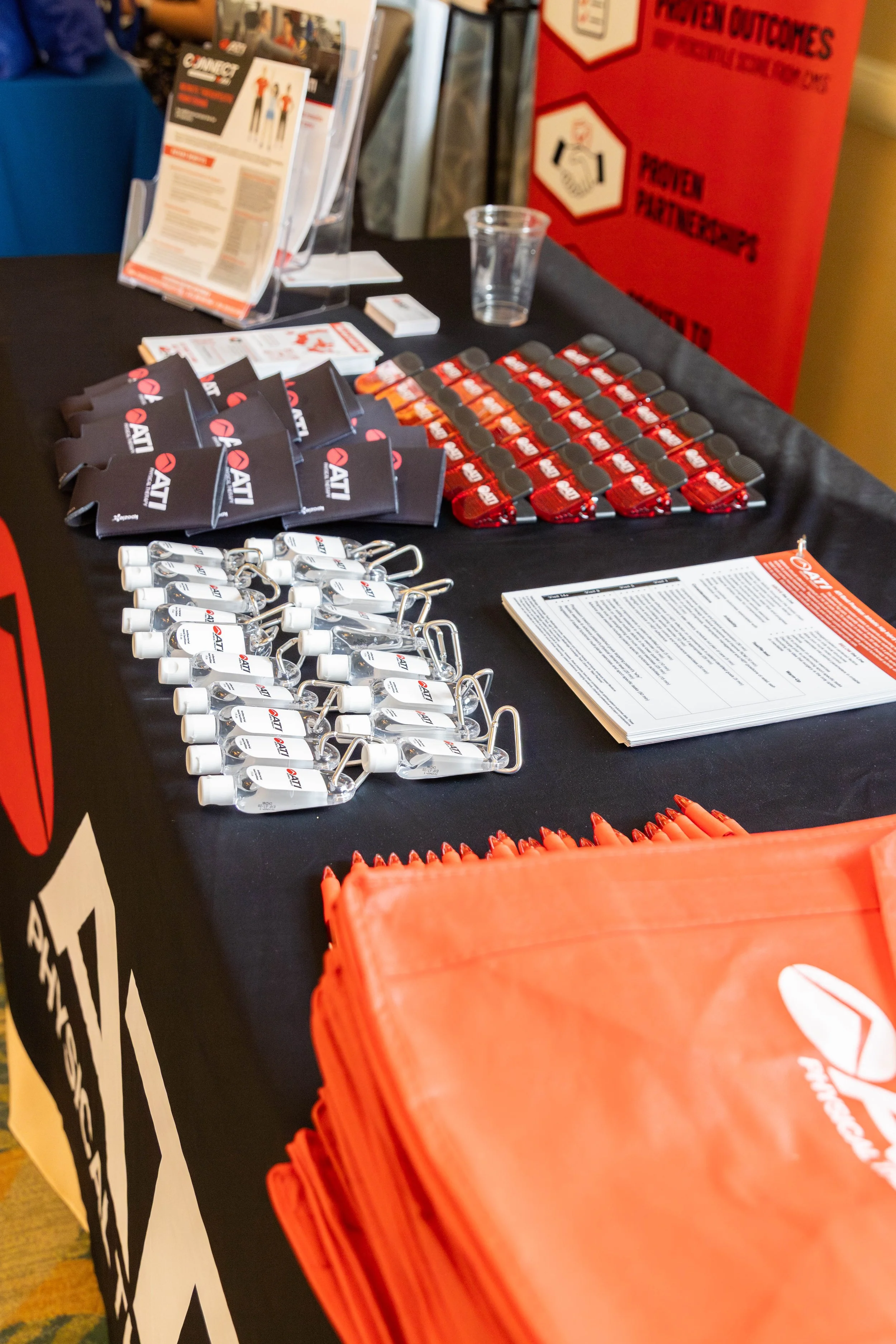 Table with ATI branded promotional materials, including small palm-sized bottles, orange lanyards, and informational leaflets, with a fire extinguisher signage in the background.