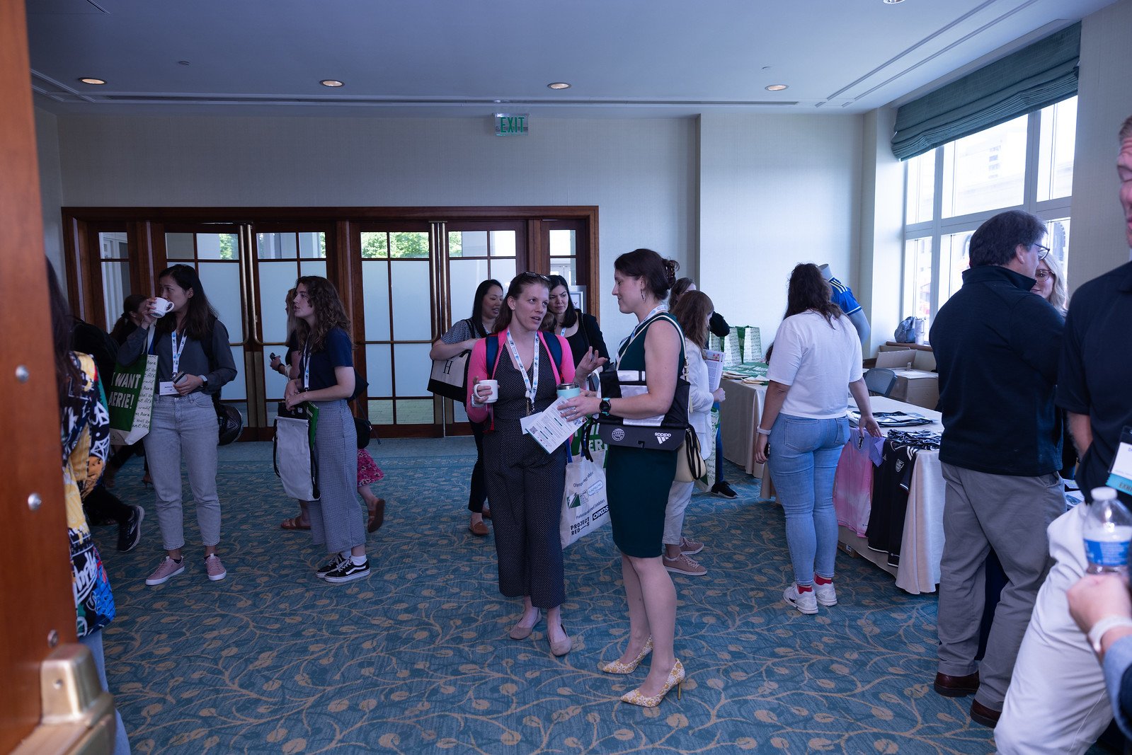 People at a conference talking and browsing booths in a well-lit room with large windows and carpeted floor.