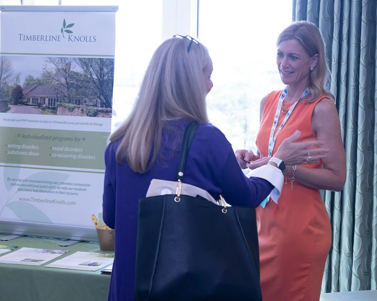 Two women having a conversation at a health or wellness conference. One woman is wearing a purple blazer with a large black handbag, and the other woman is wearing an orange dress with a white lanyard. A banner for Timberline Knolls treatment center 
