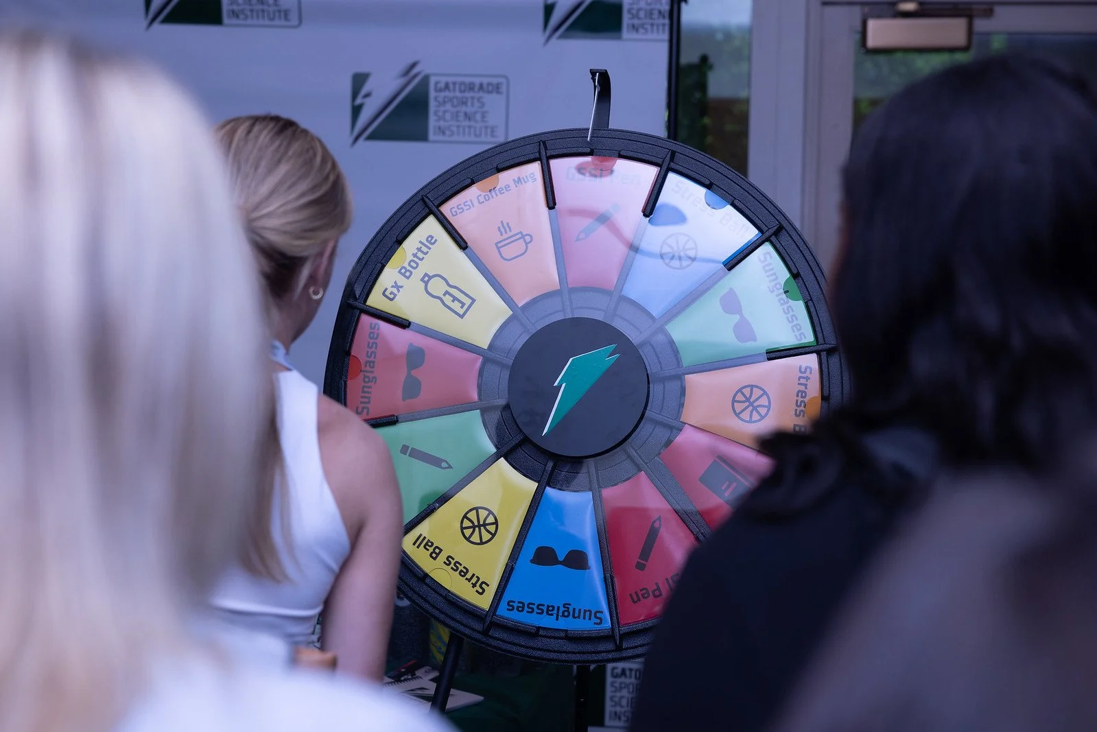People watching a spinning prize wheel with various segments, some labeled 'Sunglasses,' 'Stress Ball,' and 'GX Bottle,' at an event with 'Gatorade Sports Science Institute' logos in the background.