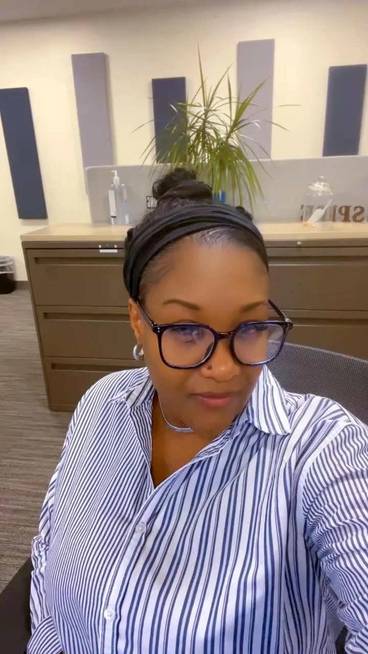 A woman with glasses and earrings taking a selfie in an office with a plant and desk in the background.