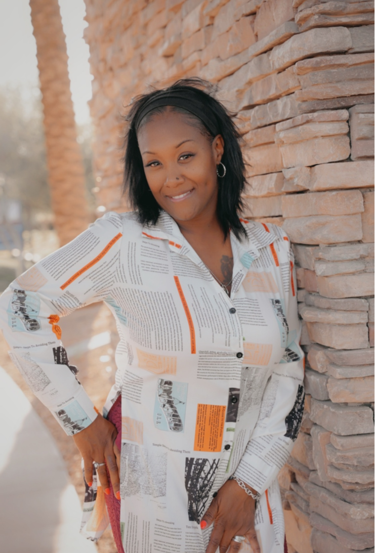A woman leaning against a stone wall outdoors, smiling at the camera. She has shoulder-length black hair, hoop earrings, and is wearing a long-sleeve button-up shirt with a newspaper print pattern.
