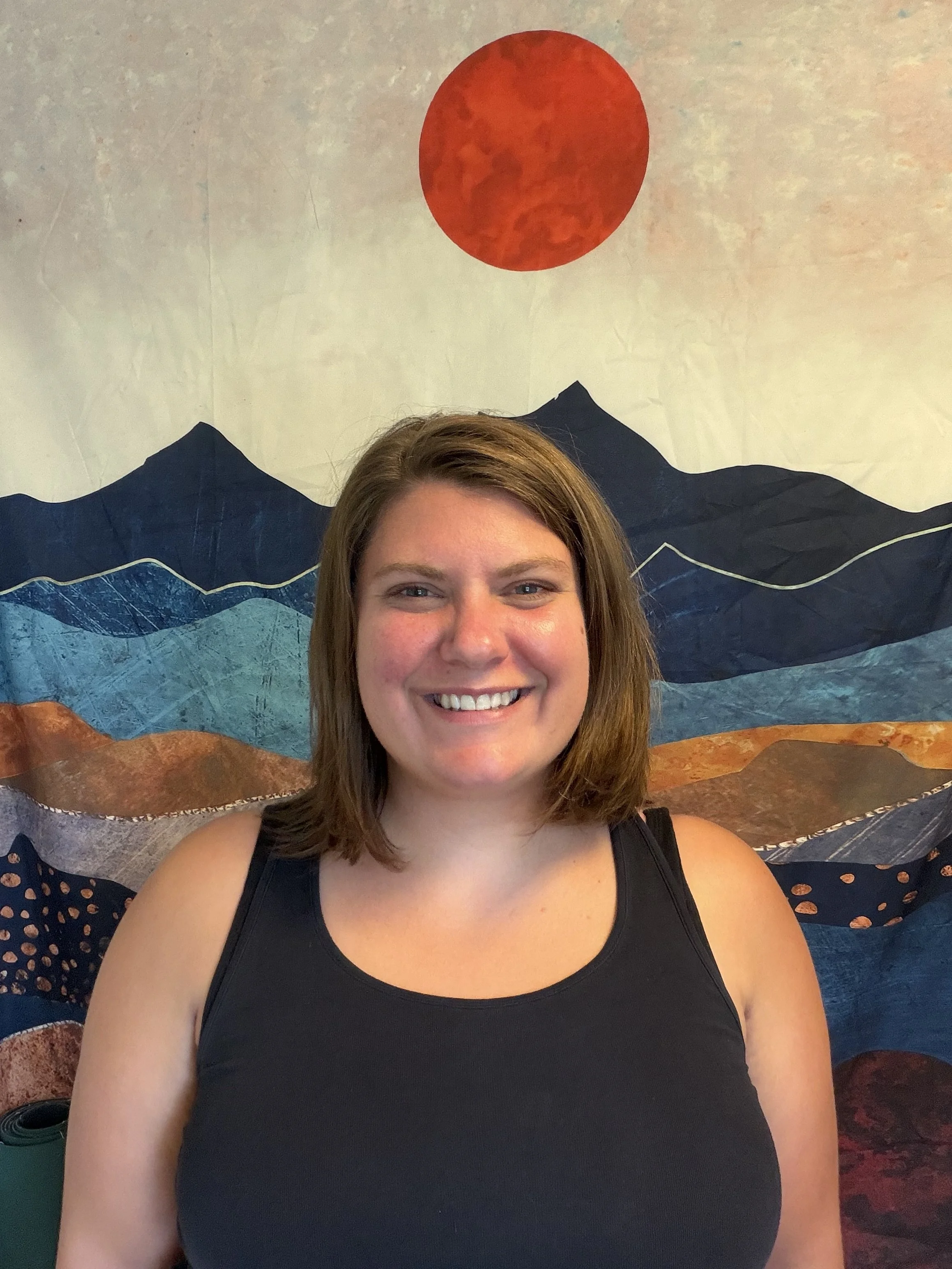 A young woman with shoulder-length brown hair, smiling, wearing a black sleeveless top, standing in front of a decorative textile wall hanging with mountain and sun motifs.