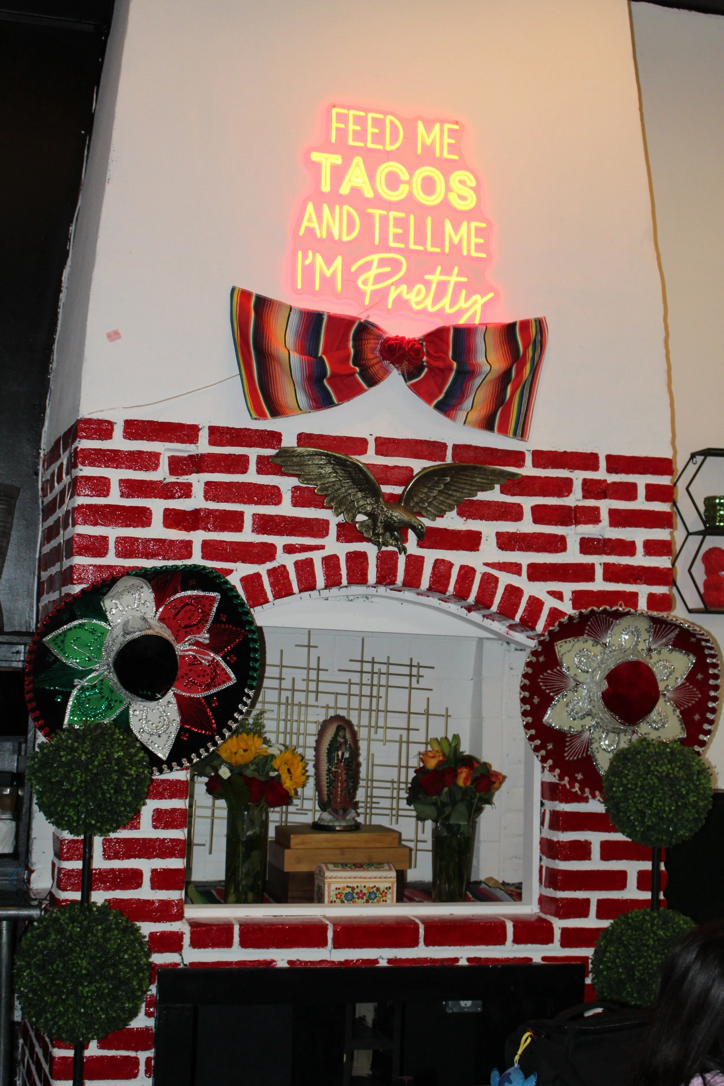 Decorative altar with red and white painted brick archway, surrounded by colorful papel picado banners, green plants, flowers, a small religious statue, and a neon sign that reads 'Feed me tacos and tell me I'm pretty'