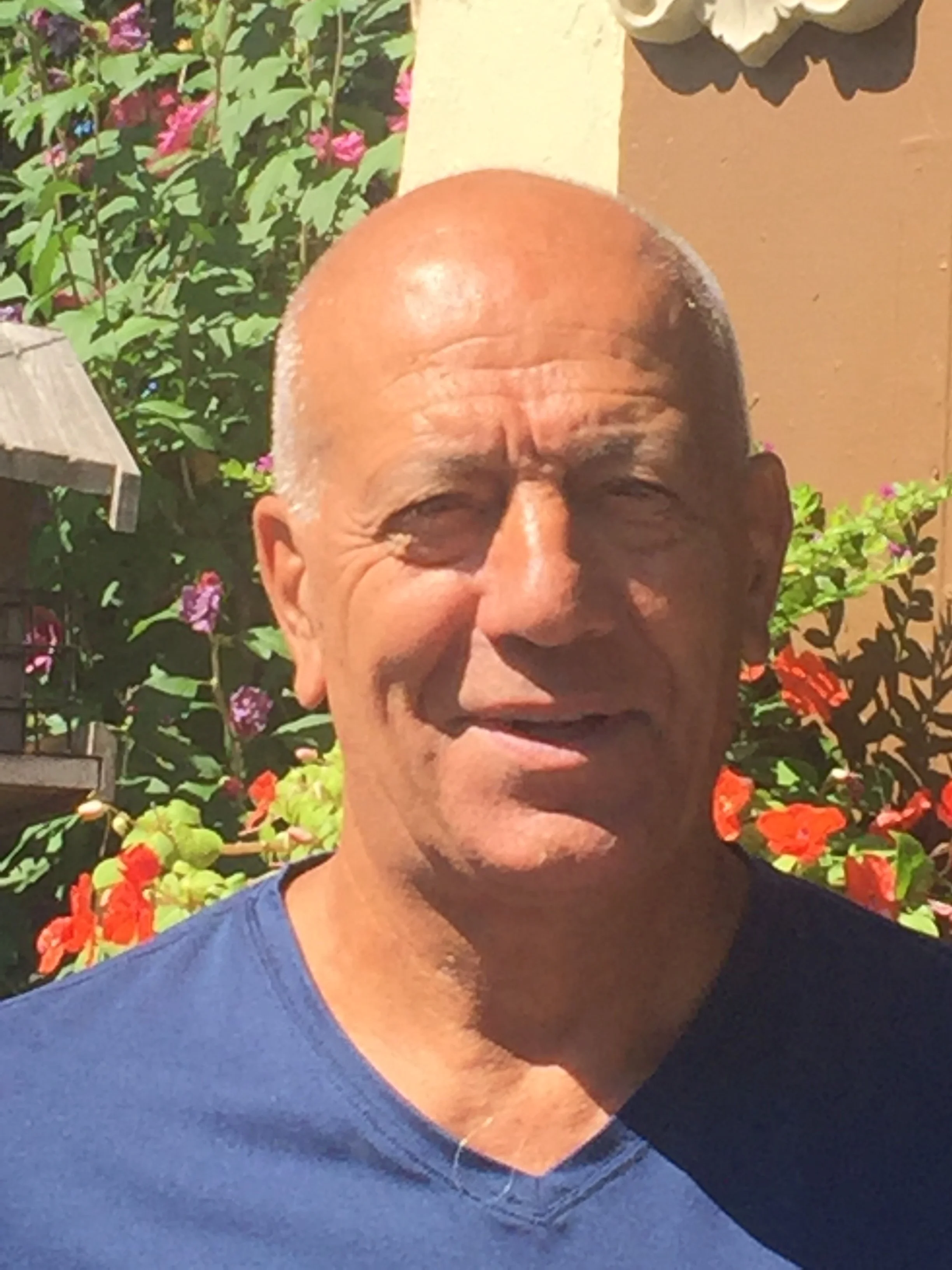 A close-up of an older man with a bald head, wearing a blue shirt, outdoors with colorful flowers and greenery in the background.