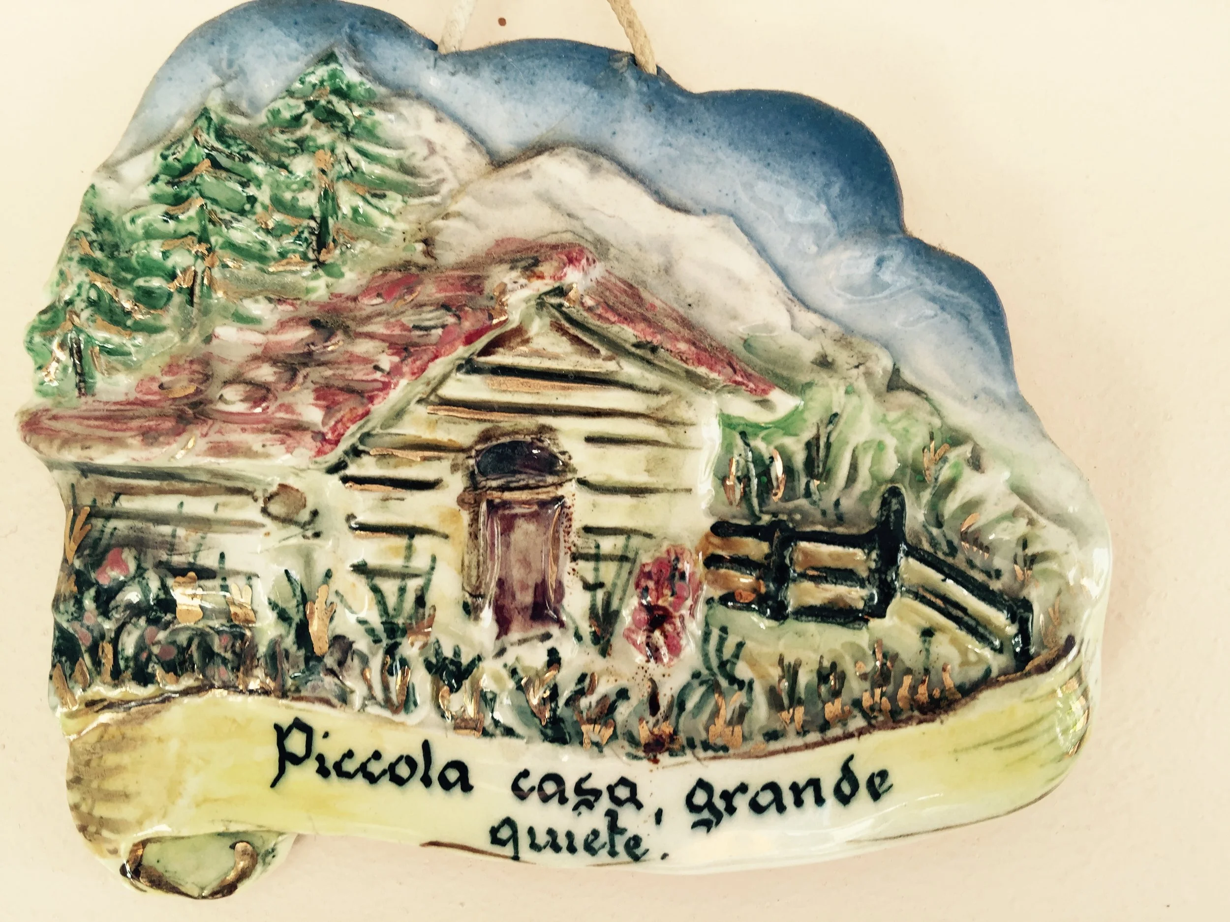 A decorative ceramic piece depicting a landscape with trees, mountains, a house, and a road, with handwritten Italian text below.