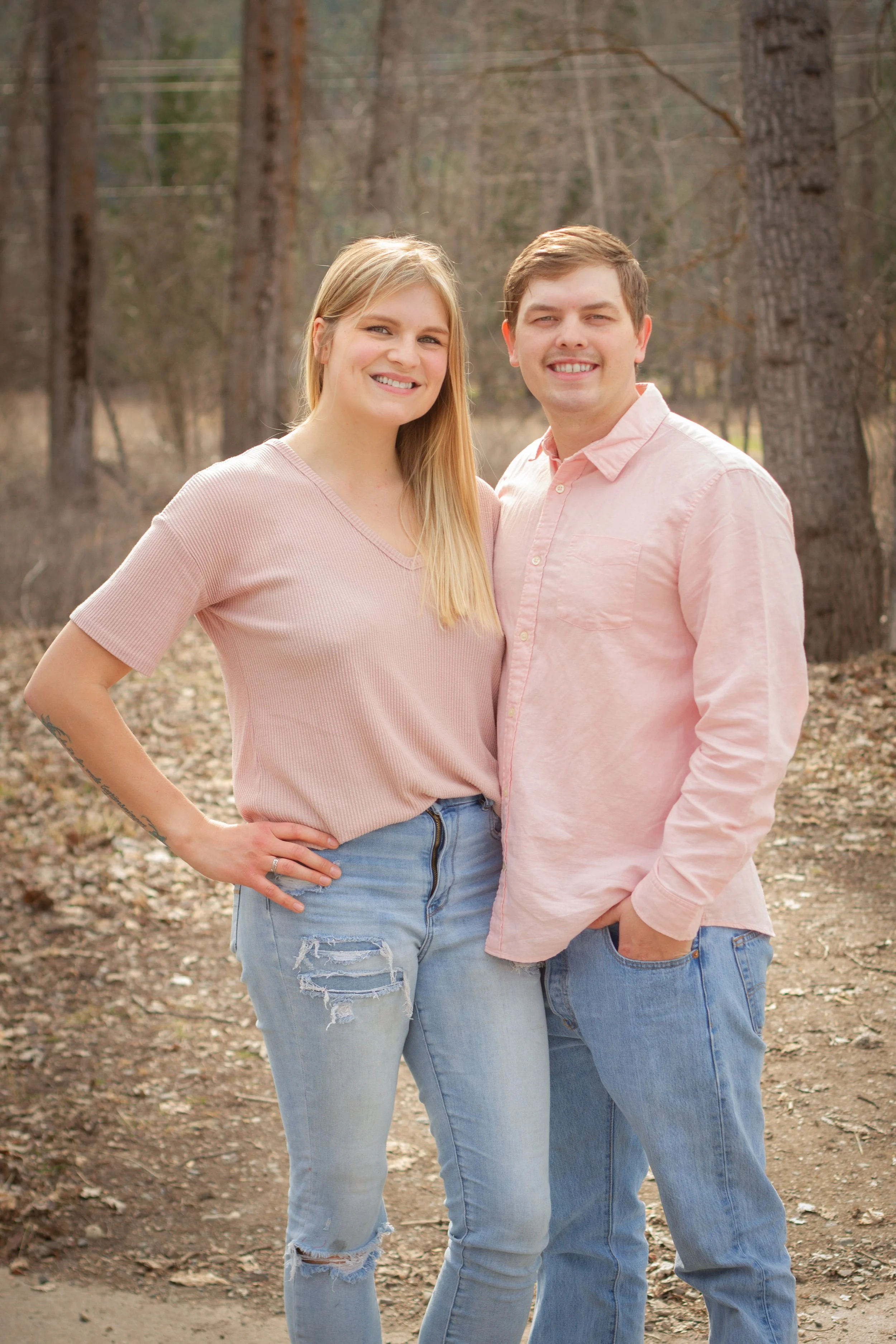 A smiling young woman and man standing close together outdoors in a wooded area with bare trees, wearing casual light-colored clothes.