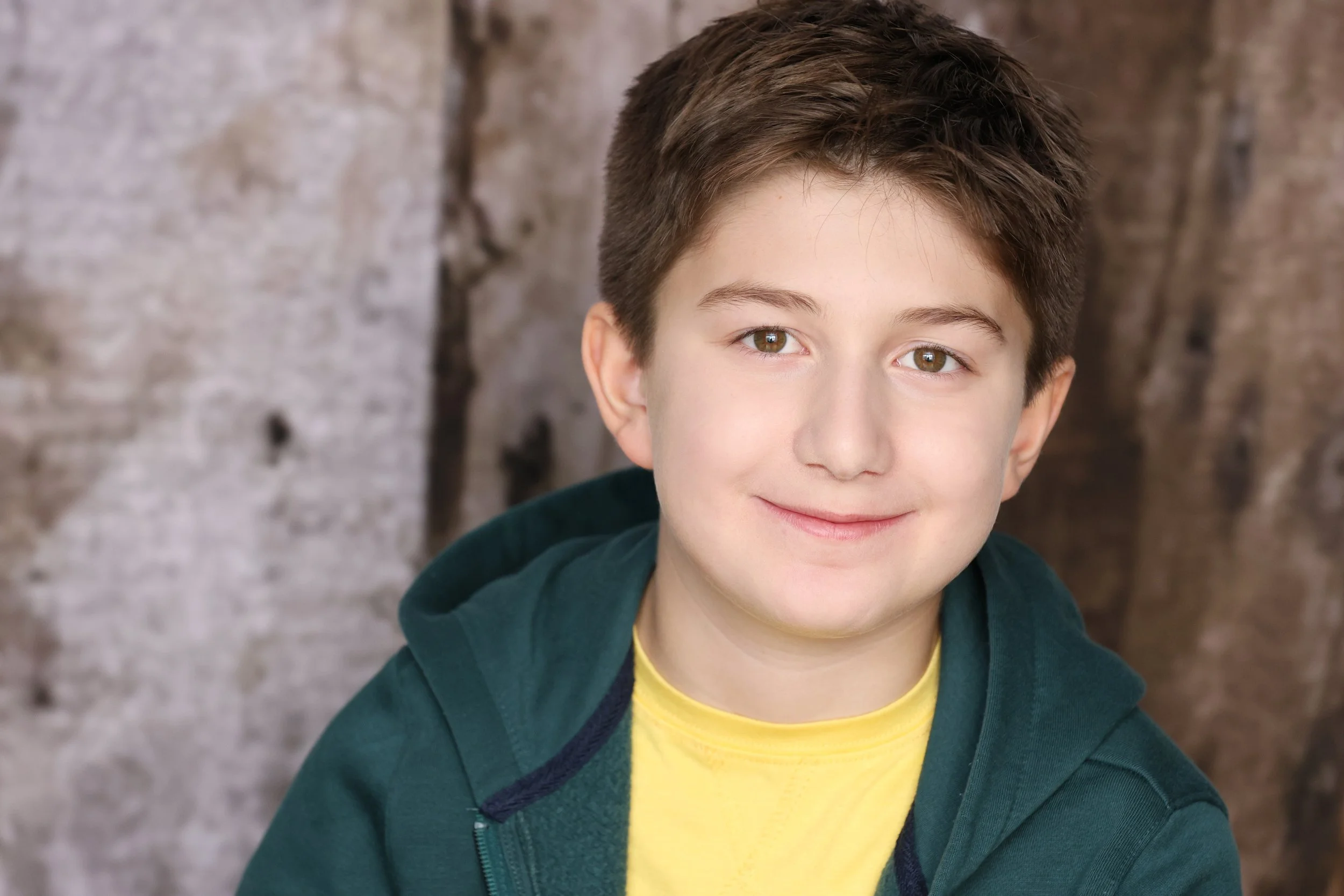 A young boy with brown hair and brown eyes smiling at the camera, wearing a yellow shirt and a dark green hoodie, standing in front of a textured stone wall.