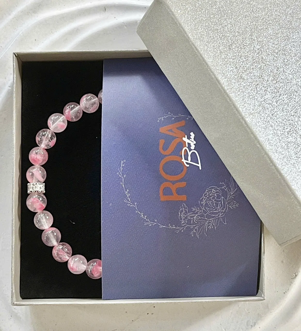 A pink beaded bracelet with a rectangular clasp inside an open jewelry box. The jewelry box has a round silver lid and a dark interior, with a blue card displaying the text "ROSA B" and floral artwork.