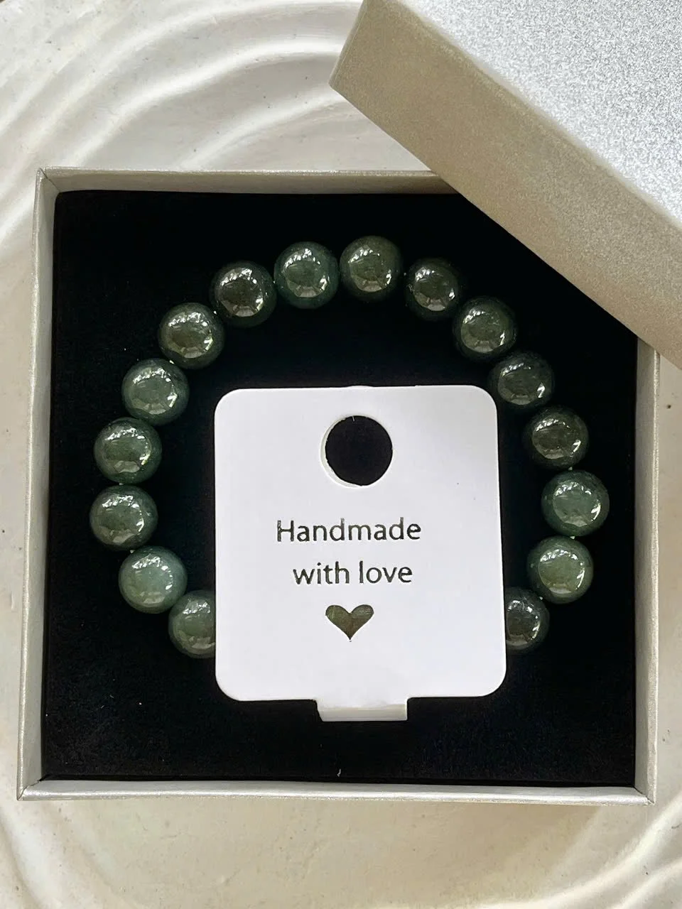 A beaded bracelet with greenish-gray beads in a jewelry box. The box has a white lid and black interior with a white tag that reads 'Handmade with love' and a small heart symbol.