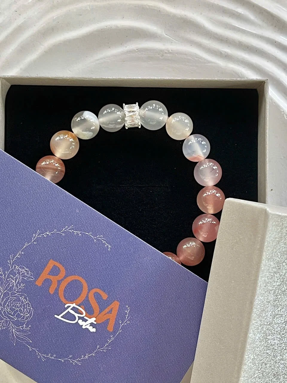 A beaded bracelet with alternating cream and pinkish beads, some with a pearlescent finish, and a silver rectangular clasp, next to a purple card with white text that reads 'ROSA But' partially covering the bracelet, all in a white jewelry box.