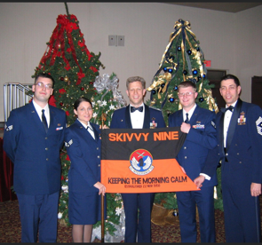 Five individuals in Air Force uniforms posing with a Christmas tree in the background and holding an orange banner that reads "SKYVY NINE KEEPING THE MORNING CALM."