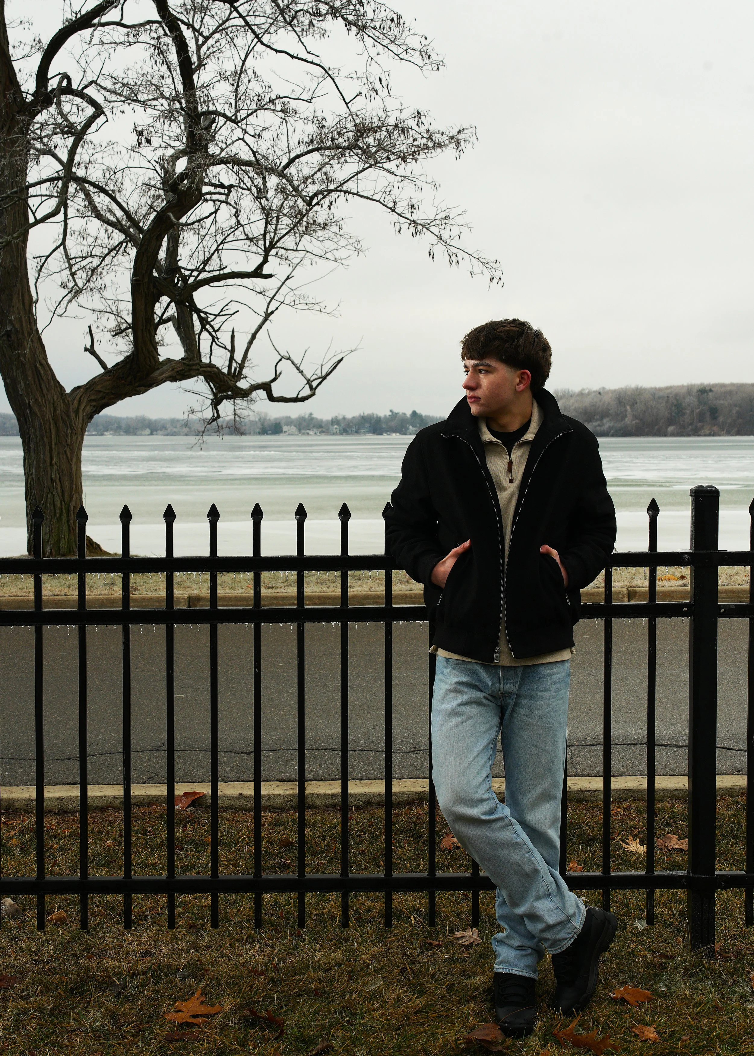 Young man standing outdoors leaning against a black fence near a frozen lake, with a leafless tree and fallen leaves on the ground under an overcast sky.