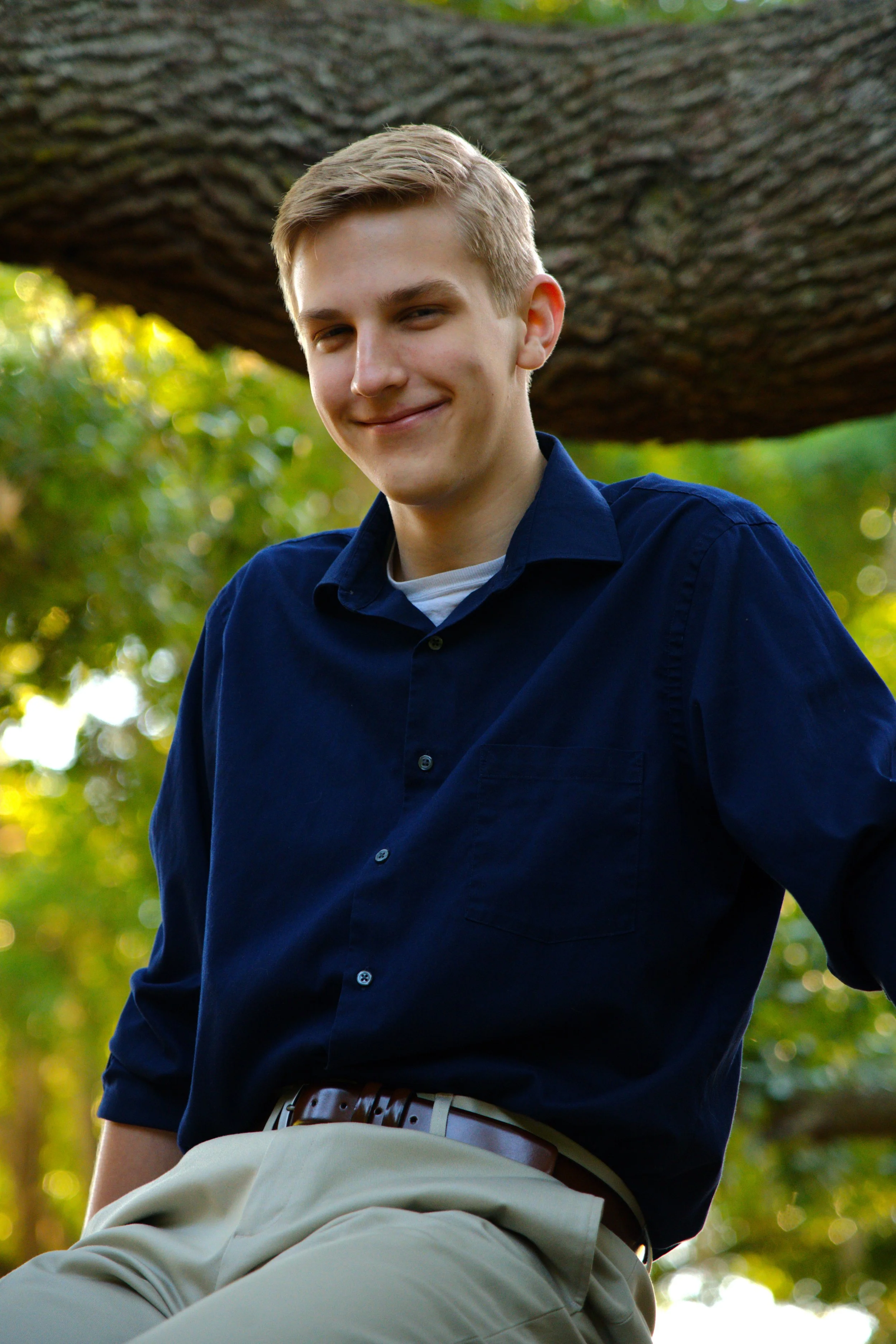 A young man with blonde hair, wearing a navy blue shirt and beige pants, sitting outdoors on a large tree with green foliage. He is smiling slightly and looking at the camera.