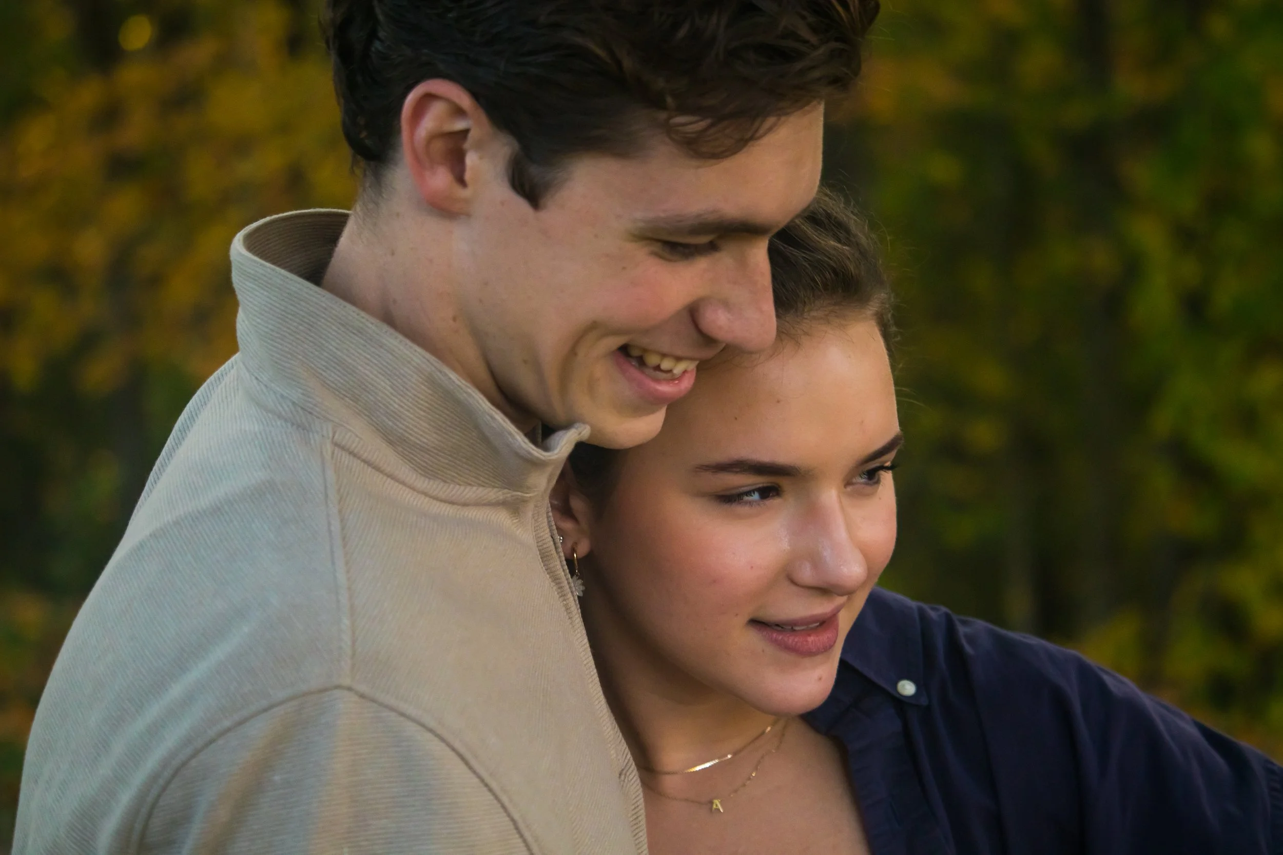 A young man and woman outside, close together and smiling, with the man gently resting his forehead against the woman's head. Autumn trees with yellow and green leaves are in the background.