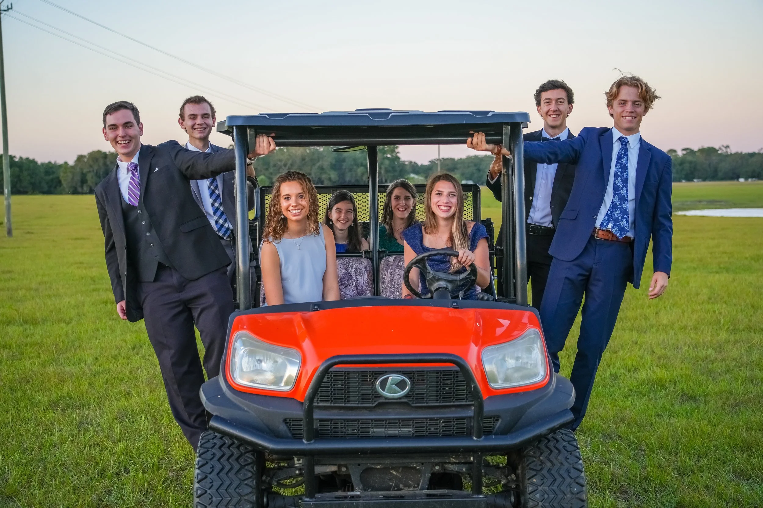 Group of young people in suits riding and standing on an RTV in a grassy field.