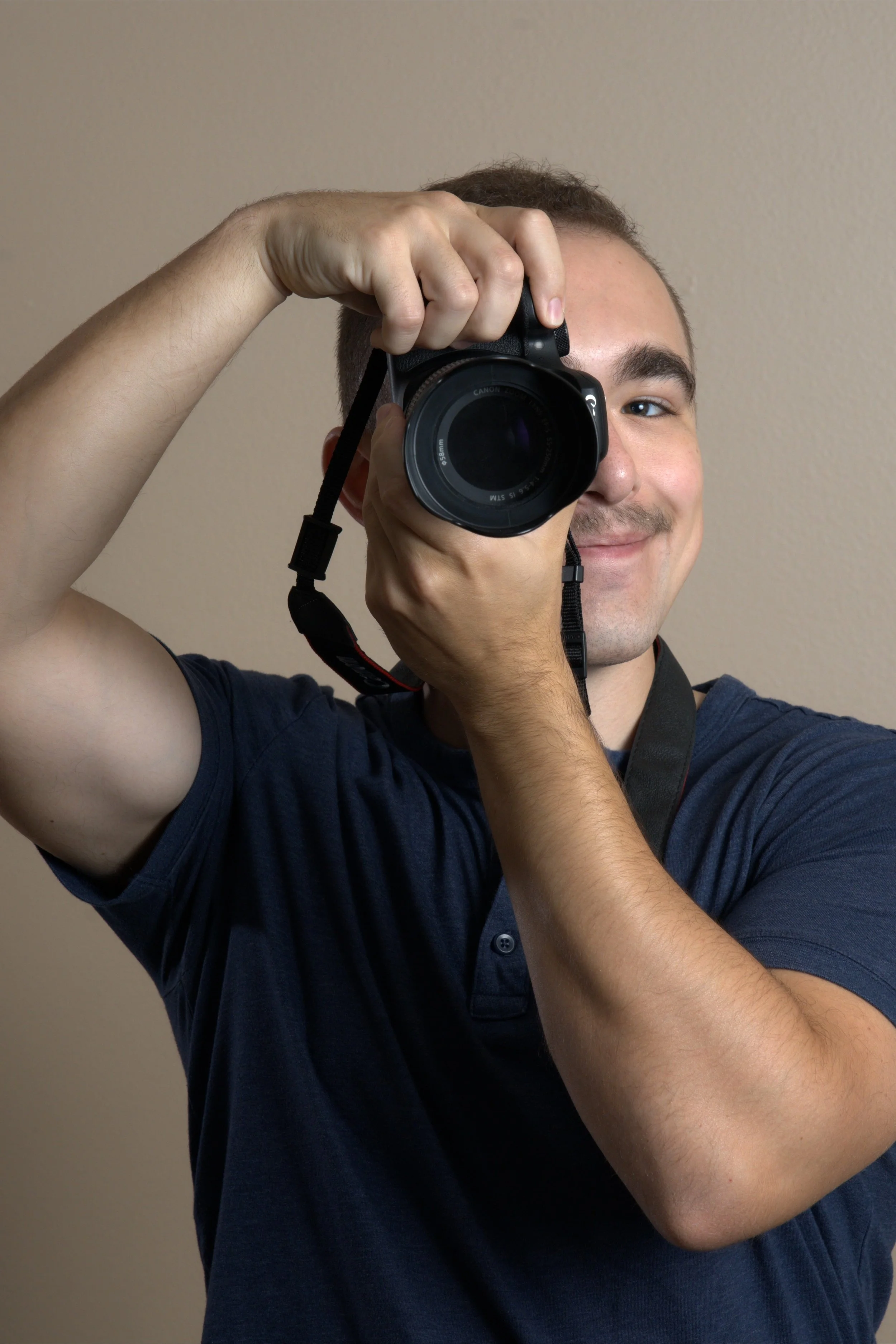 A young man with short hair and a slight smile is looking through a camera, which he is holding up to his face. He is wearing a navy blue shirt and standing in front of a plain brown background.