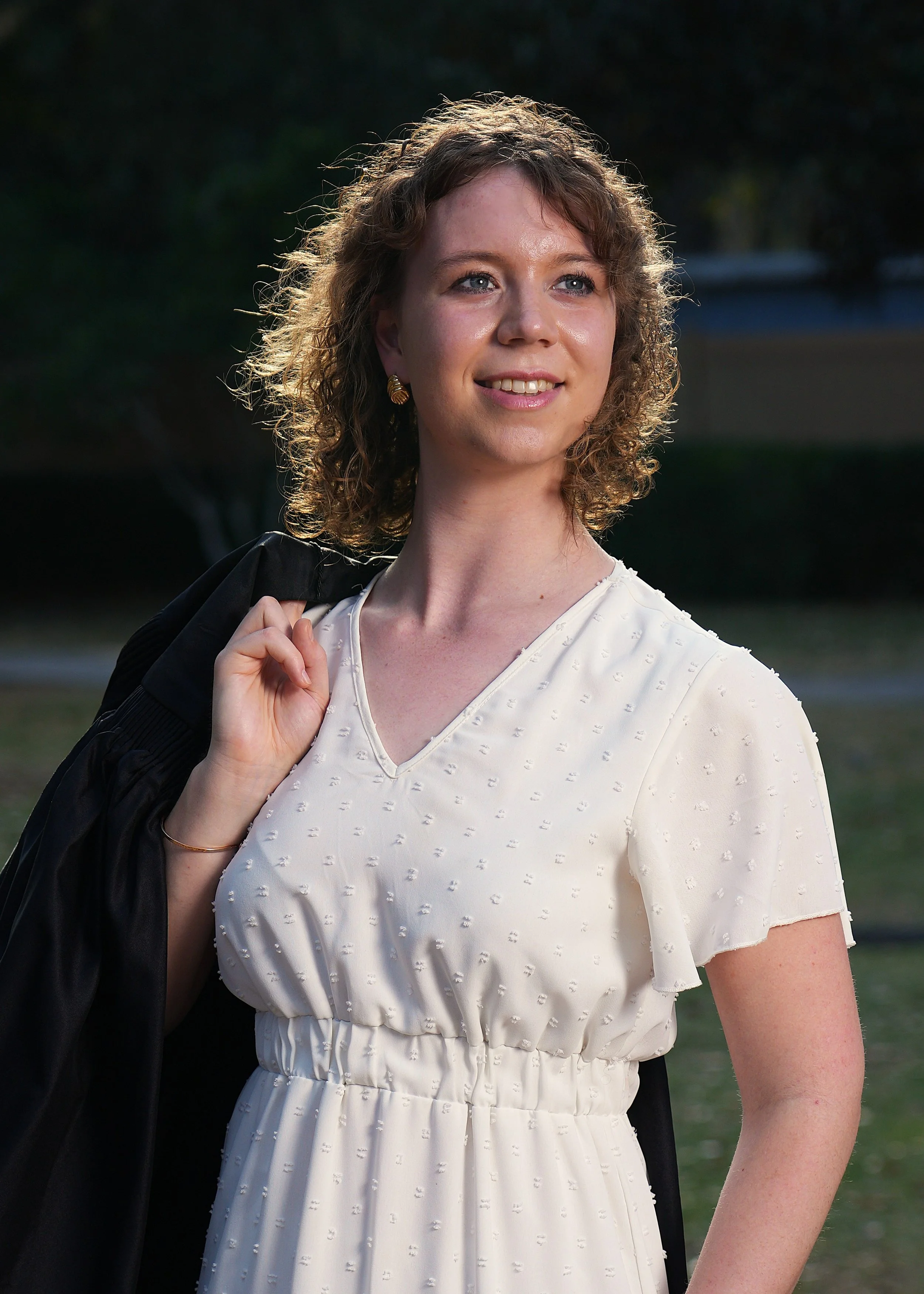 A young woman outdoors, smiling, with curly dark blonde hair, wearing a white dress with a textured pattern and a ruffled sleeve, a black graduation gown thrown over one shoulder.