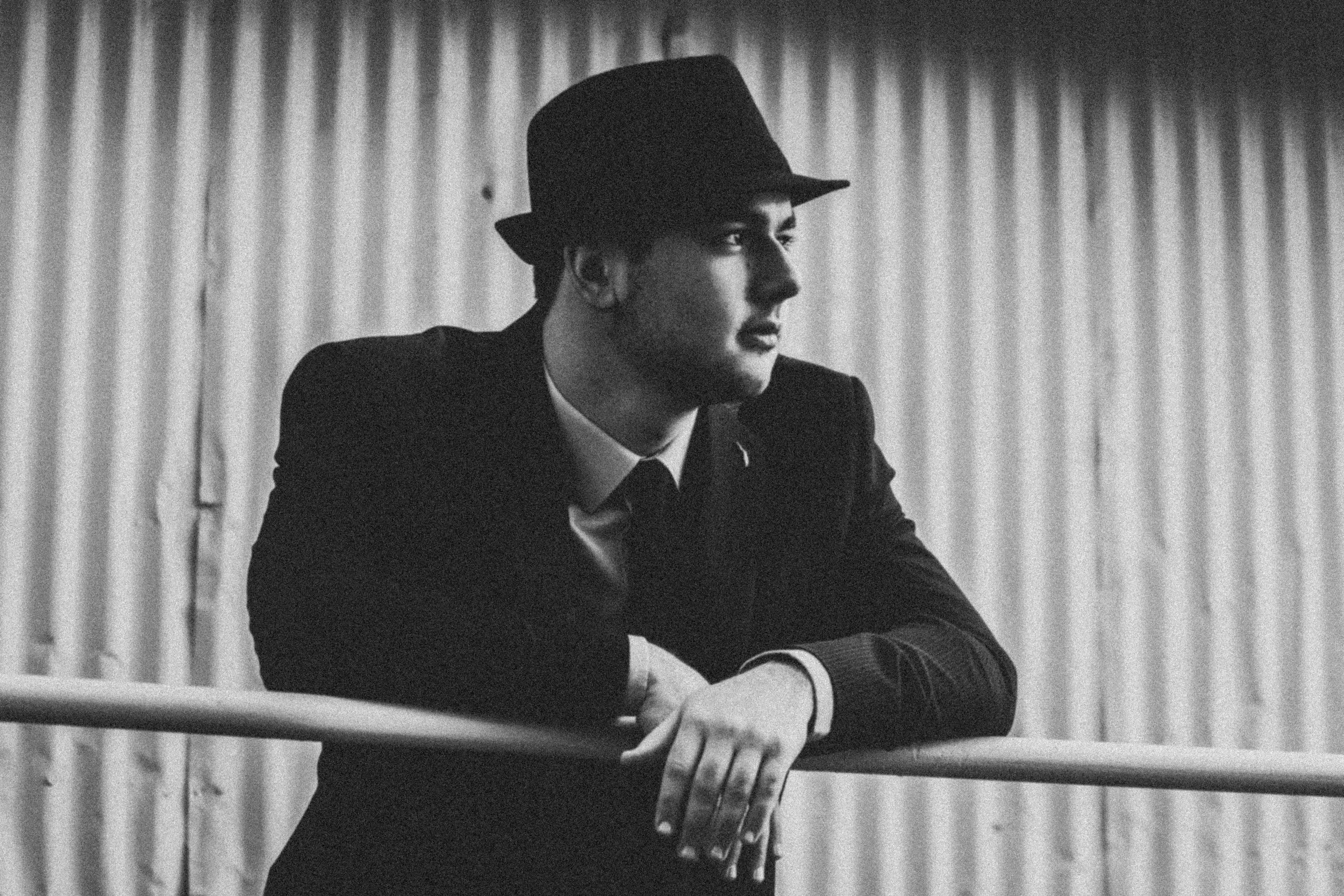 A black and white photo of a man wearing a fedora and a suit, leaning his elbows on a metal railing and looking off to one side.
