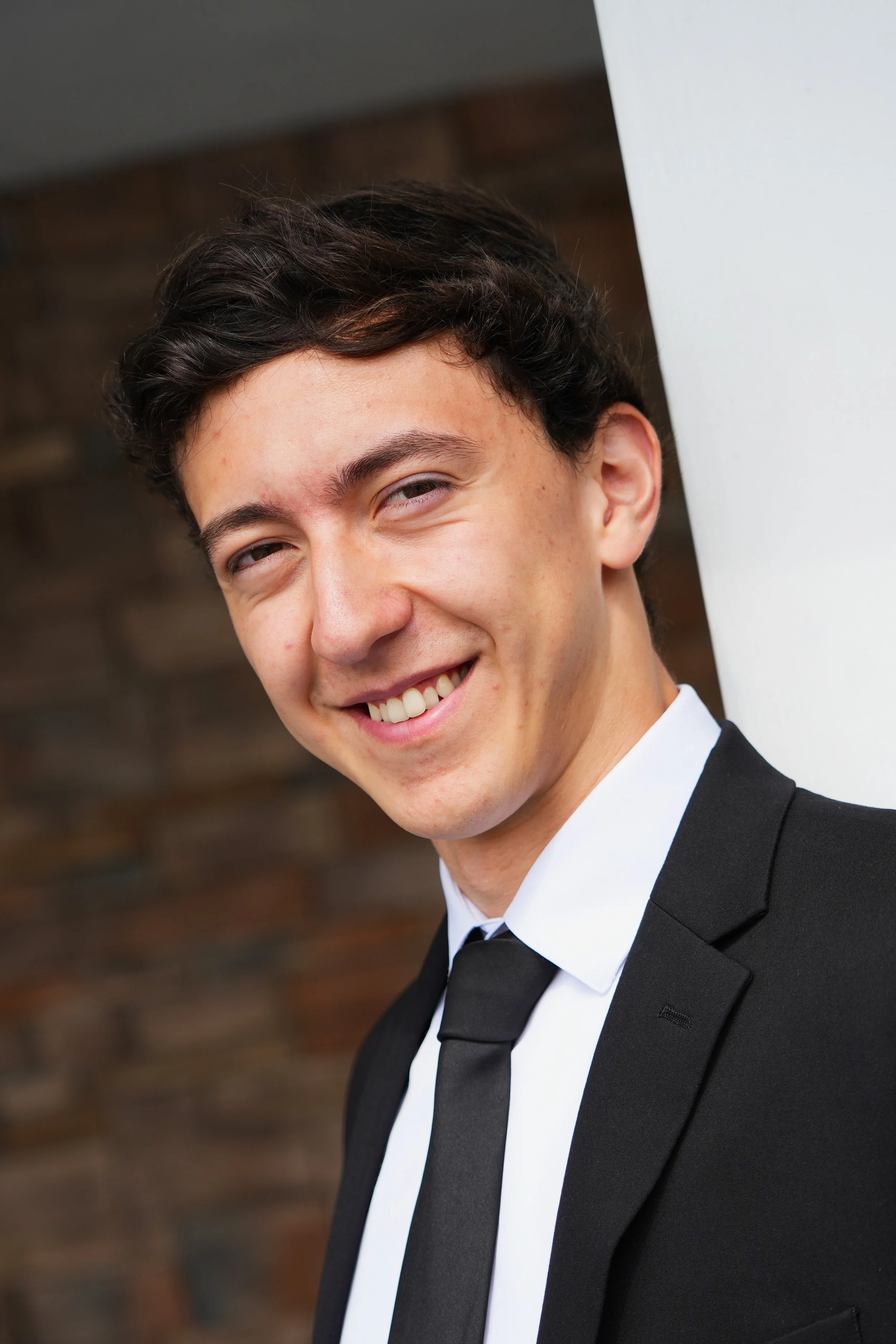 Close-up of a young man smiling in formal attire, wearing a black suit with a white shirt and black tie, against a brown brick background.