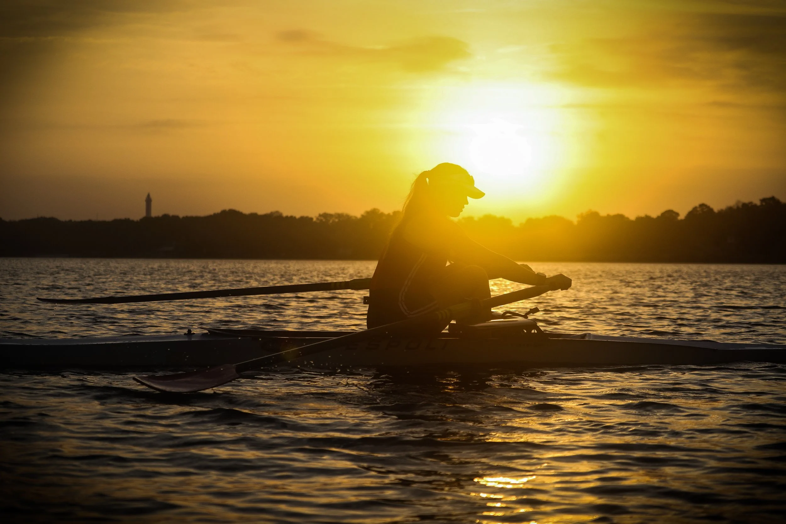 Silhouette of a person rowing a boat on a lake at sunrise, with the sun just appearing over a distant shore with a lone tower in the background.