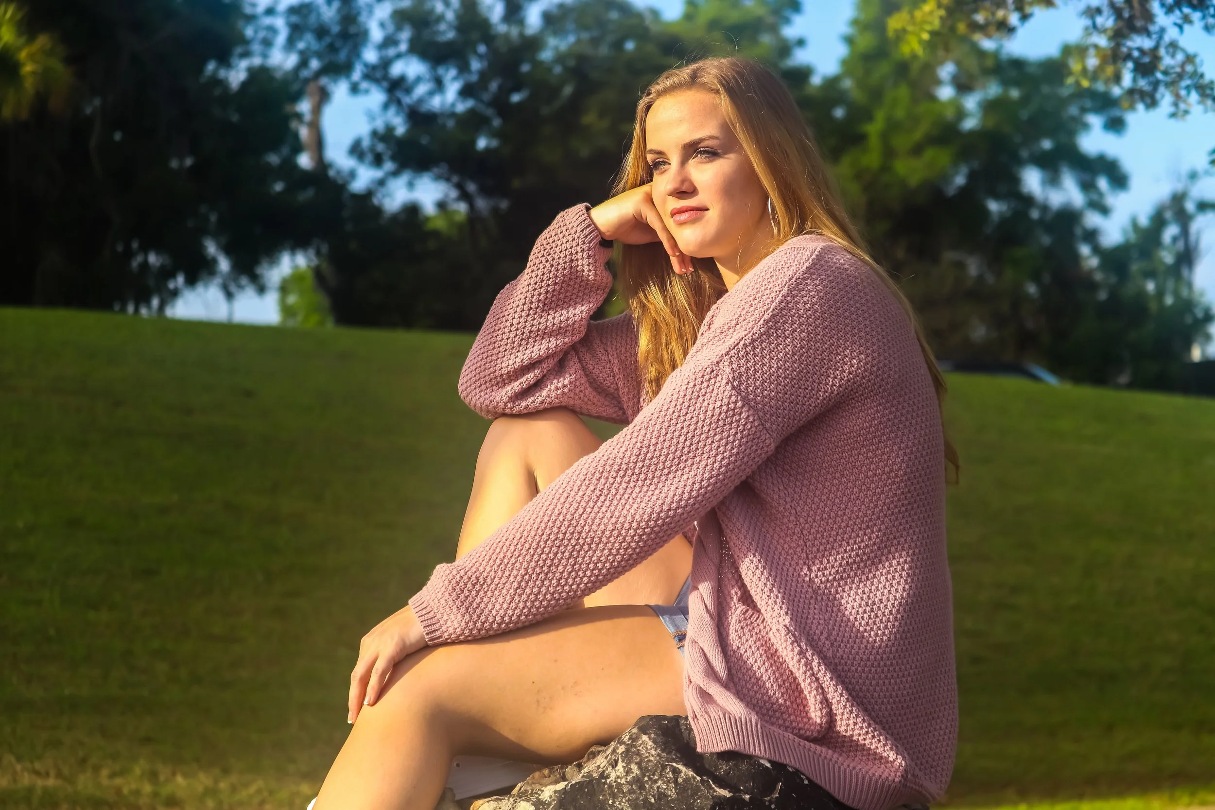 A woman sitting outdoors on a rock, with a pink sweater draped over her shoulders, looking thoughtfully to the side with a backdrop of green trees and blue sky.