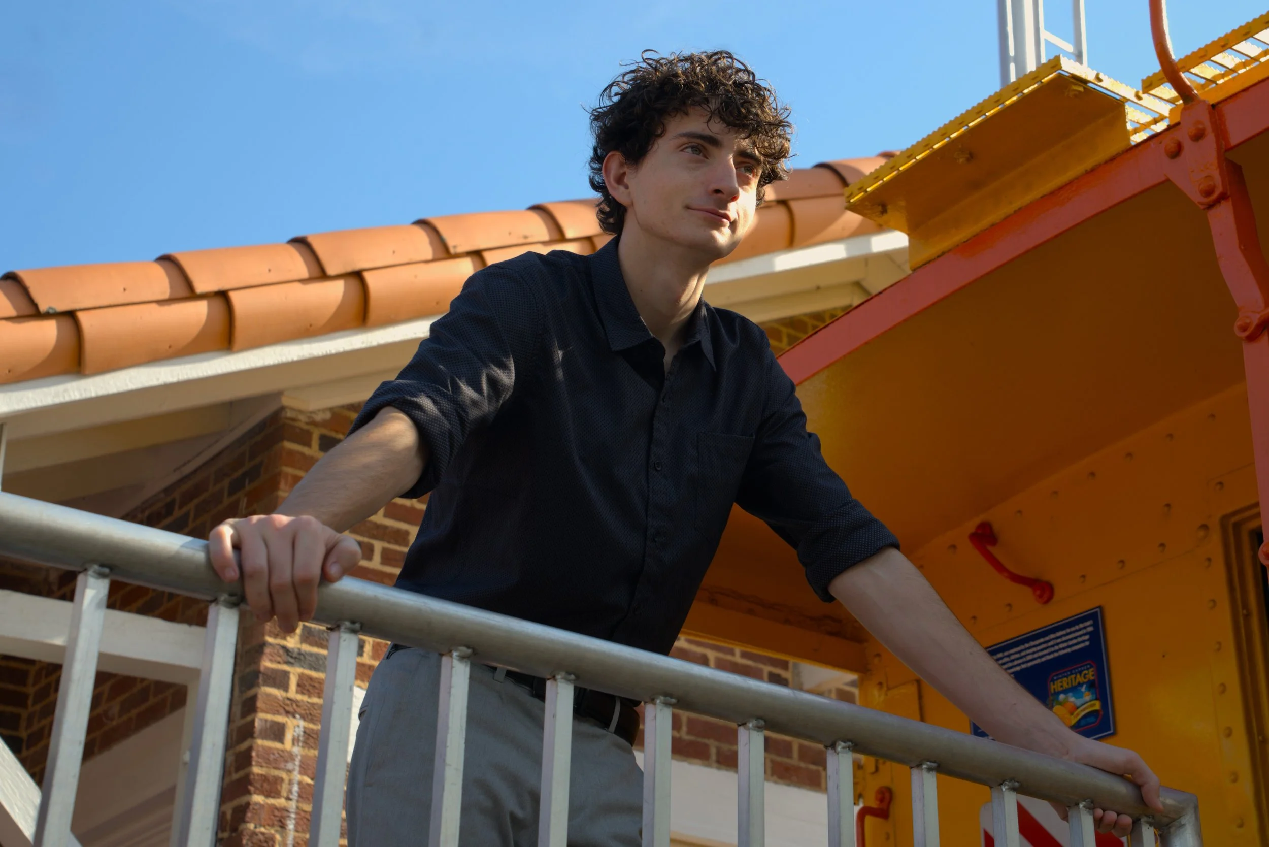 A young man with curly dark hair and wearing a black shirt is leaning on a white railing outdoors, with a brick building and a yellow train car in the background, under a clear blue sky.