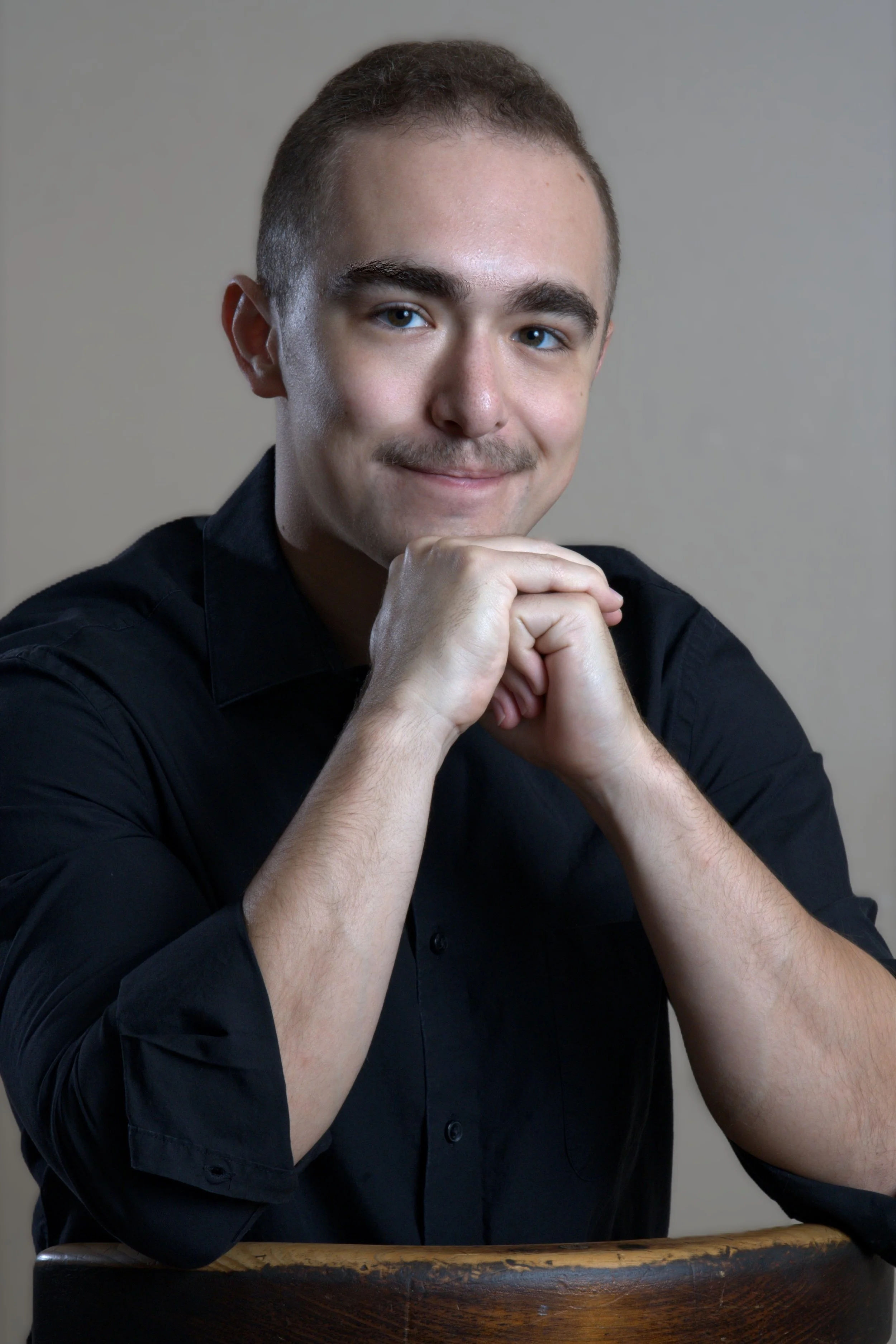 A young man with short brown hair, brown eyes, and light skin, smiling softly, resting his chin on his clasped hands, wearing a black shirt, sitting on a wooden chair against a gray background.