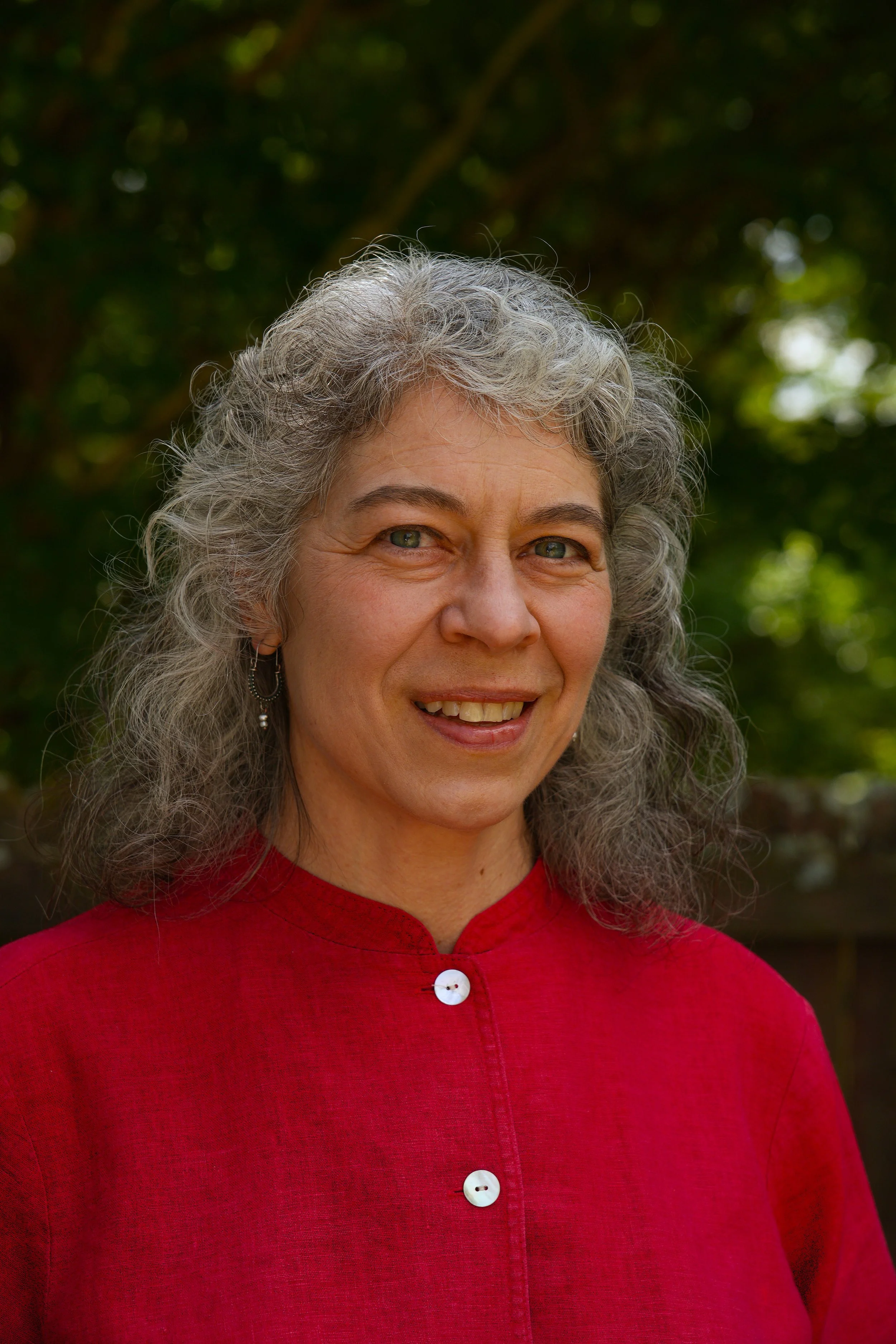 A woman with curly graying hair and dark green eyes, smiling, wearing a red top with three white buttons, standing outdoors with blurred green foliage in the background.