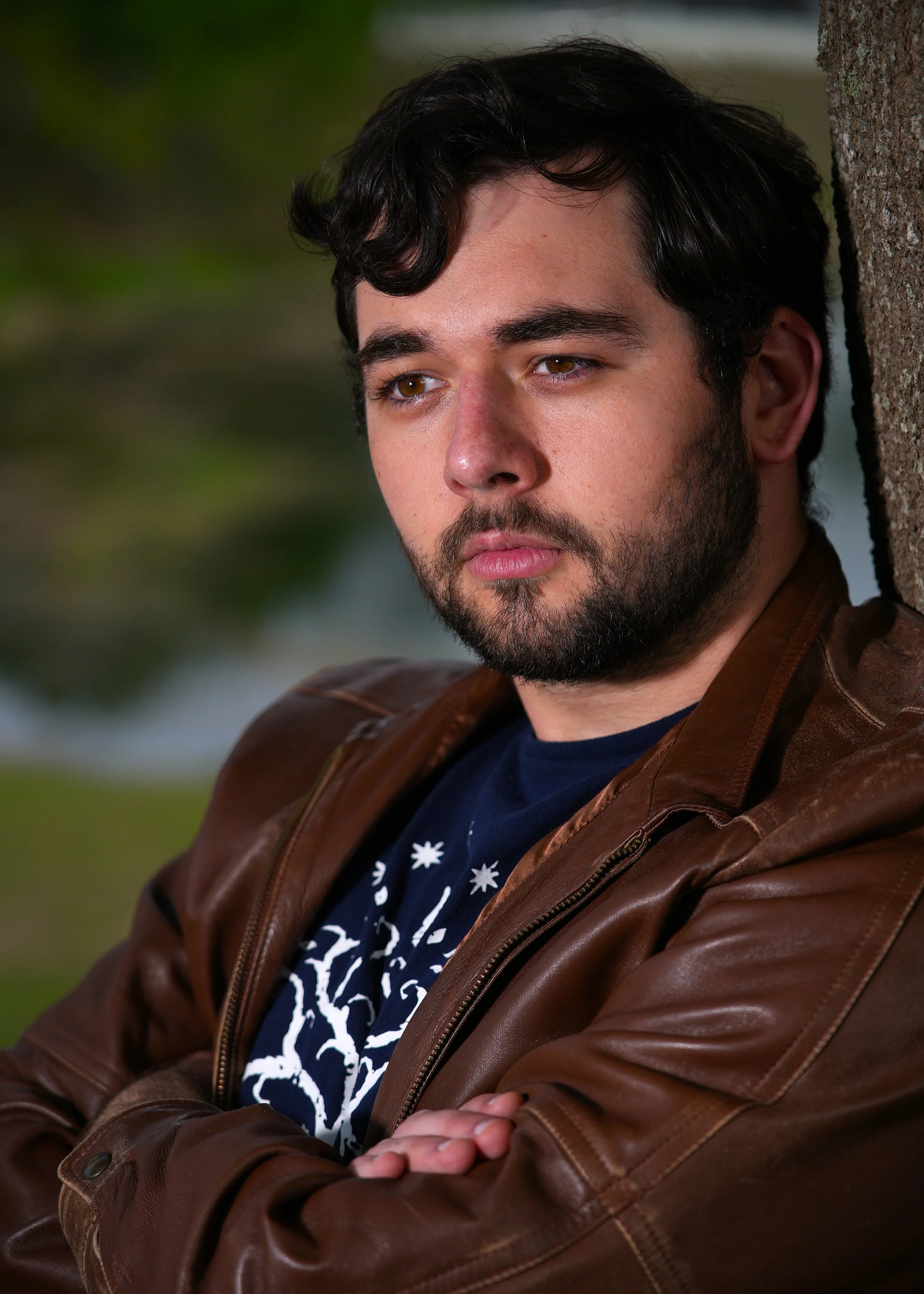 A young man with dark, wavy hair, a beard, and brown eyes is leaning against a tree with his arms crossed. He is wearing a brown leather jacket and a dark shirt with white patterns. The background is blurred with green foliage.