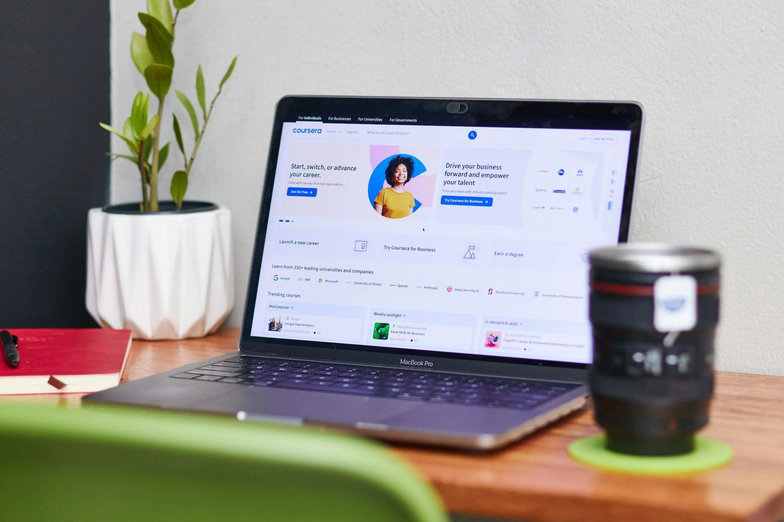 A laptop on a wooden desk displays the Coursera website. To the left of the laptop, there's a white potted plant with green leaves. In front of the laptop, items like notebooks, a pen, and a coffee cup are partially visible. The background shows a white wall.