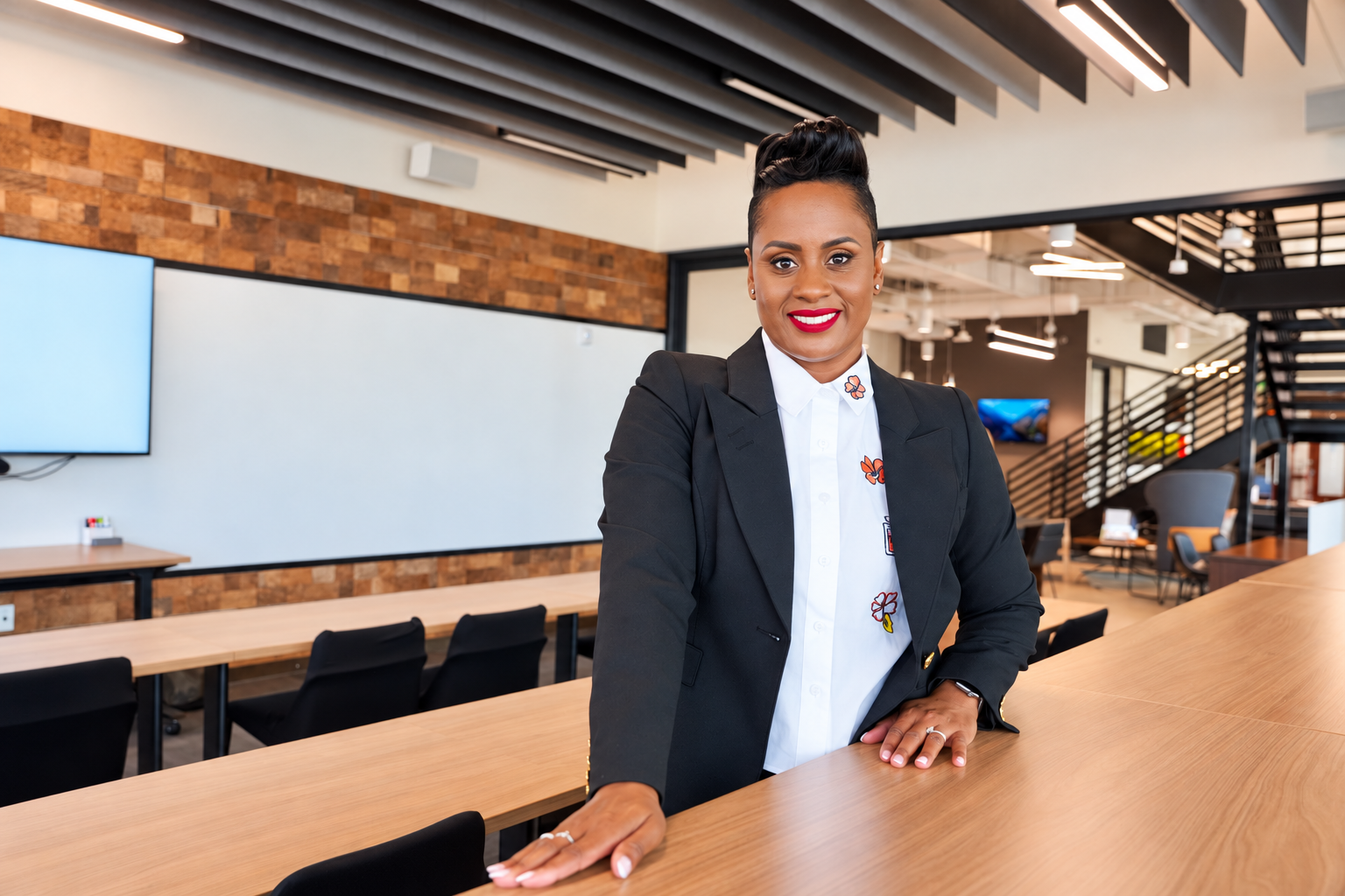 A confident woman in business attire standing in a modern conference room with wooden tables and a brick wall.