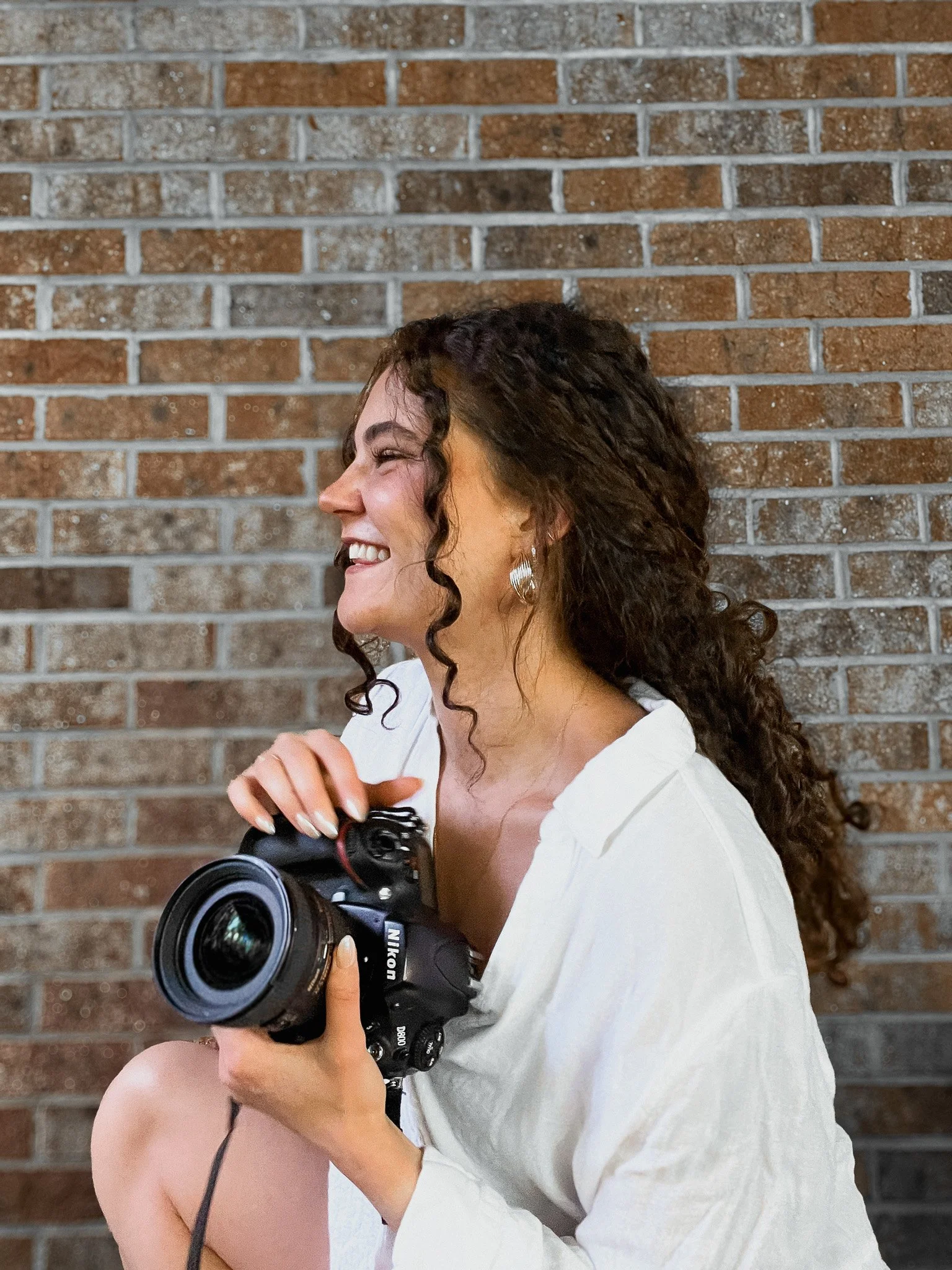 A woman with curly dark hair smiling and holding a Nikon DSLR camera, wearing a white top and silver earrings, standing against a brick wall background.