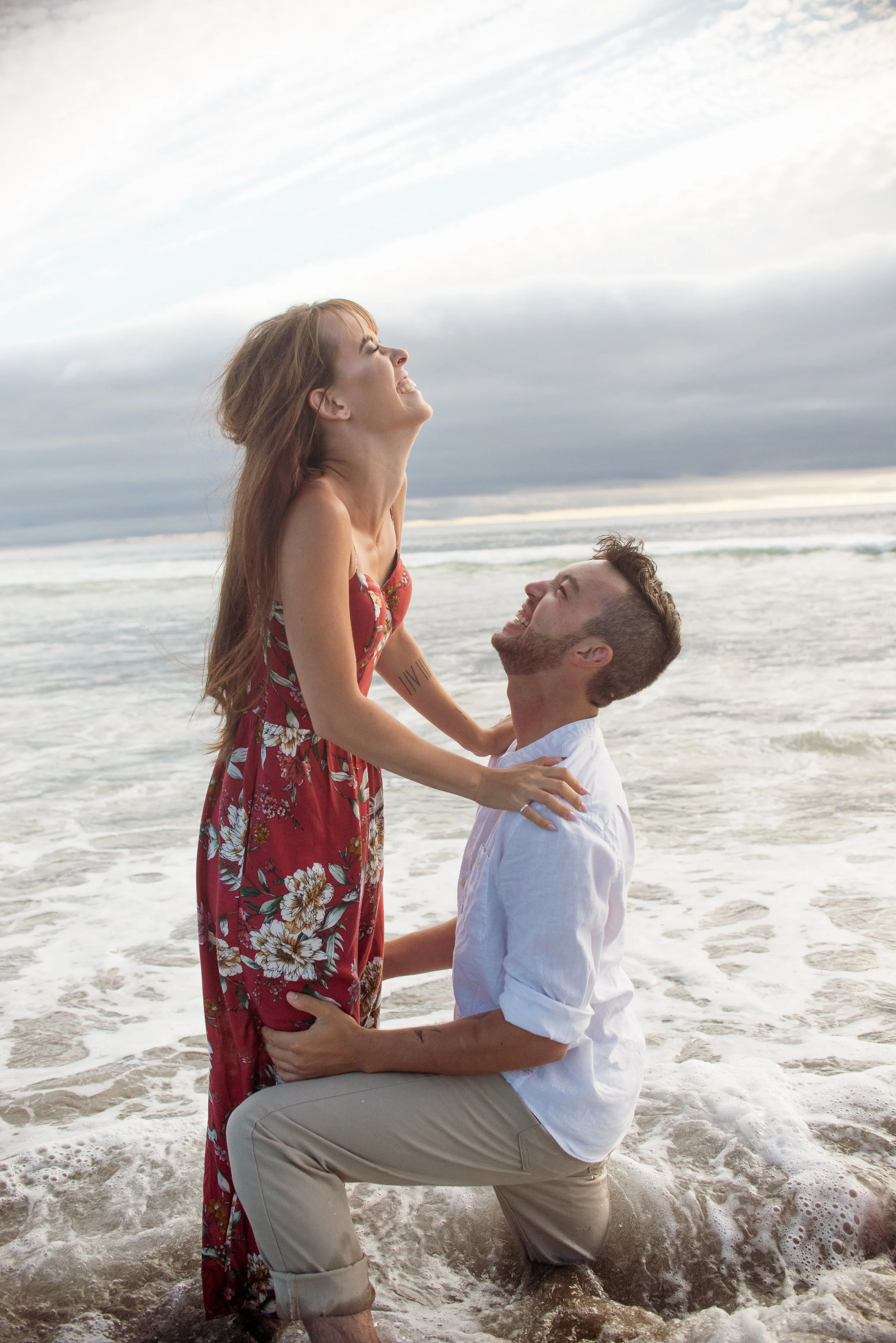 A man kneeling in the ocean holding a woman in his arms, both smiling, with a cloudy sky and the sea in the background, during a beach wedding or romantic moment.