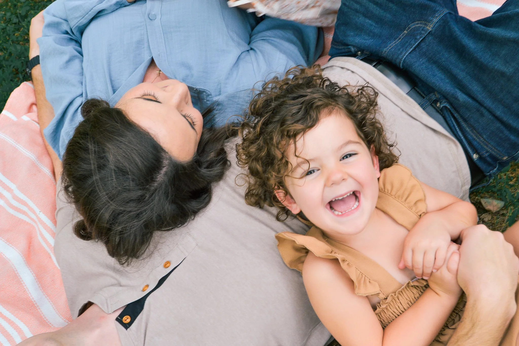A woman with dark hair is lying on her back on a blanket, with a young girl with curly hair smiling and hugging her. The woman has her eyes closed, and the girl is looking at the camera, laughing.